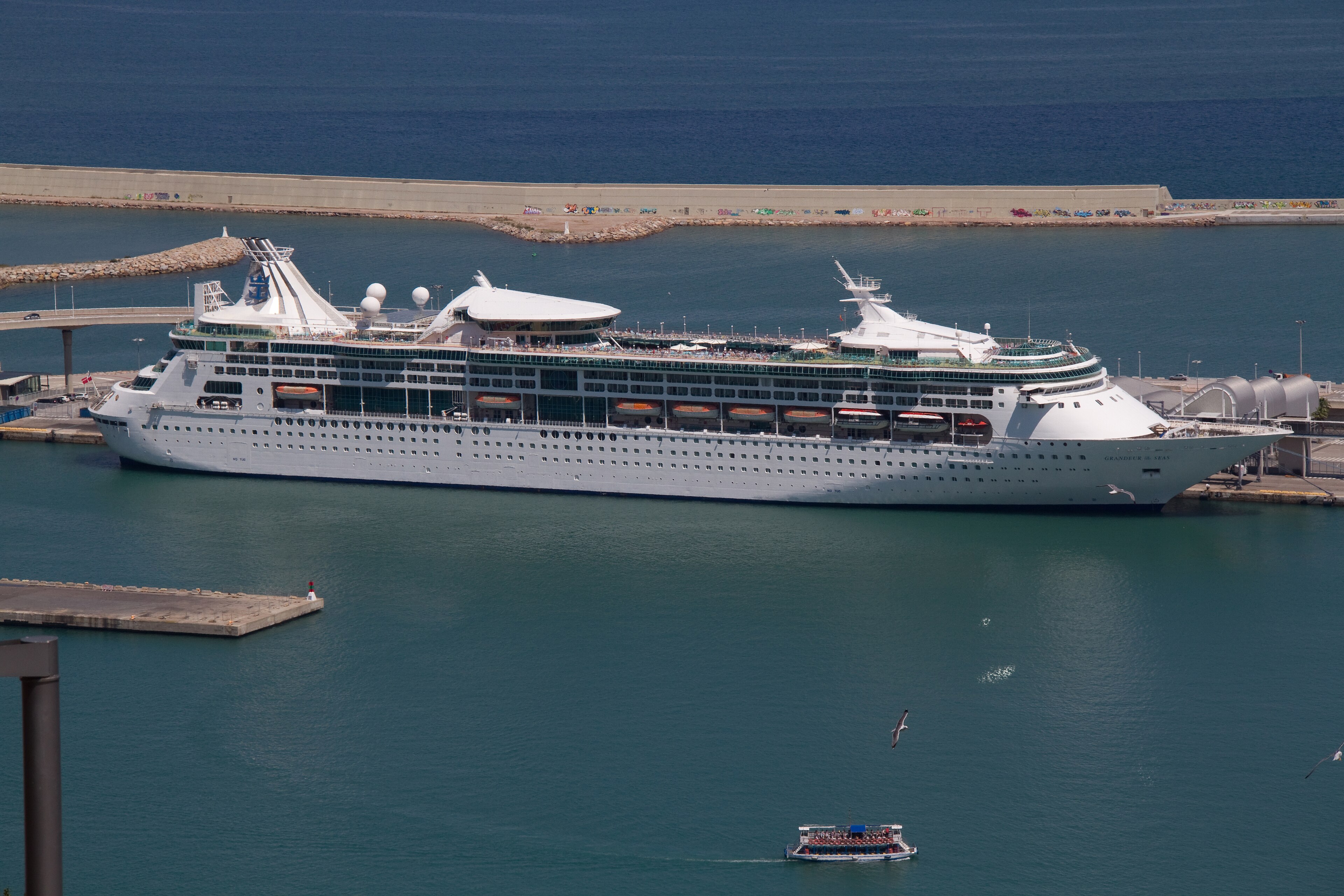 Ein großes weißes Kreuzfahrtschiff liegt im Hafen neben einer Pier, während ein kleineres Ausflugsboot auf dem Wasser fährt.