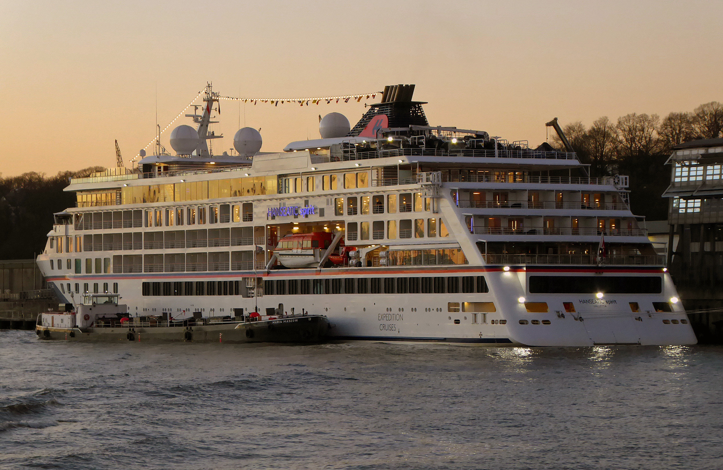 Ein großes weißes Kreuzfahrtschiff namens Hanseatic Spirit liegt im Hafen, beleuchtet von Innenlichtern und Außenstrahlern, während die Sonne untergeht.