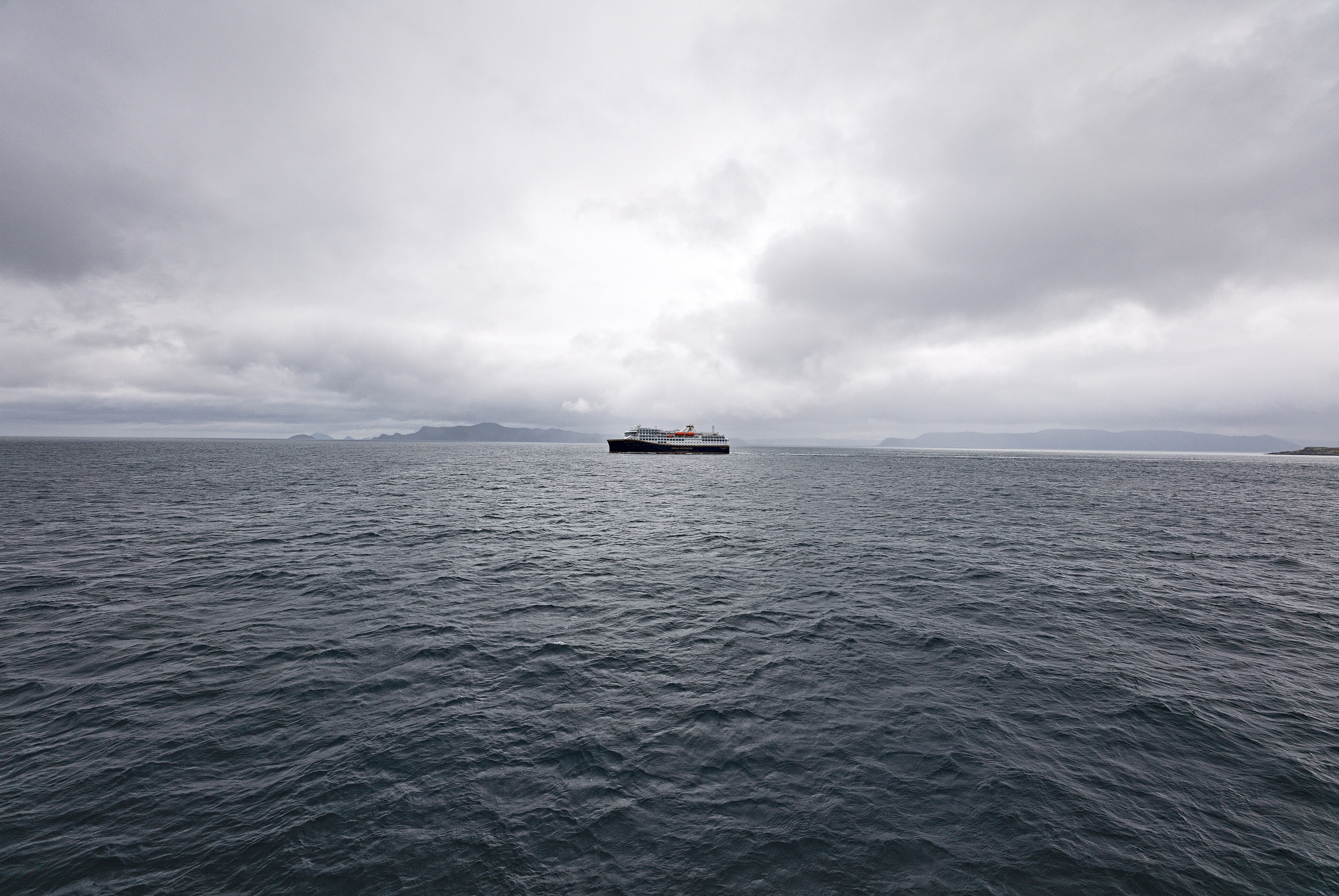 Ein großes Passagierschiff fährt auf dem Meer unter einer bewölkten Himmel, im Hintergrund sind Berge zu sehen.