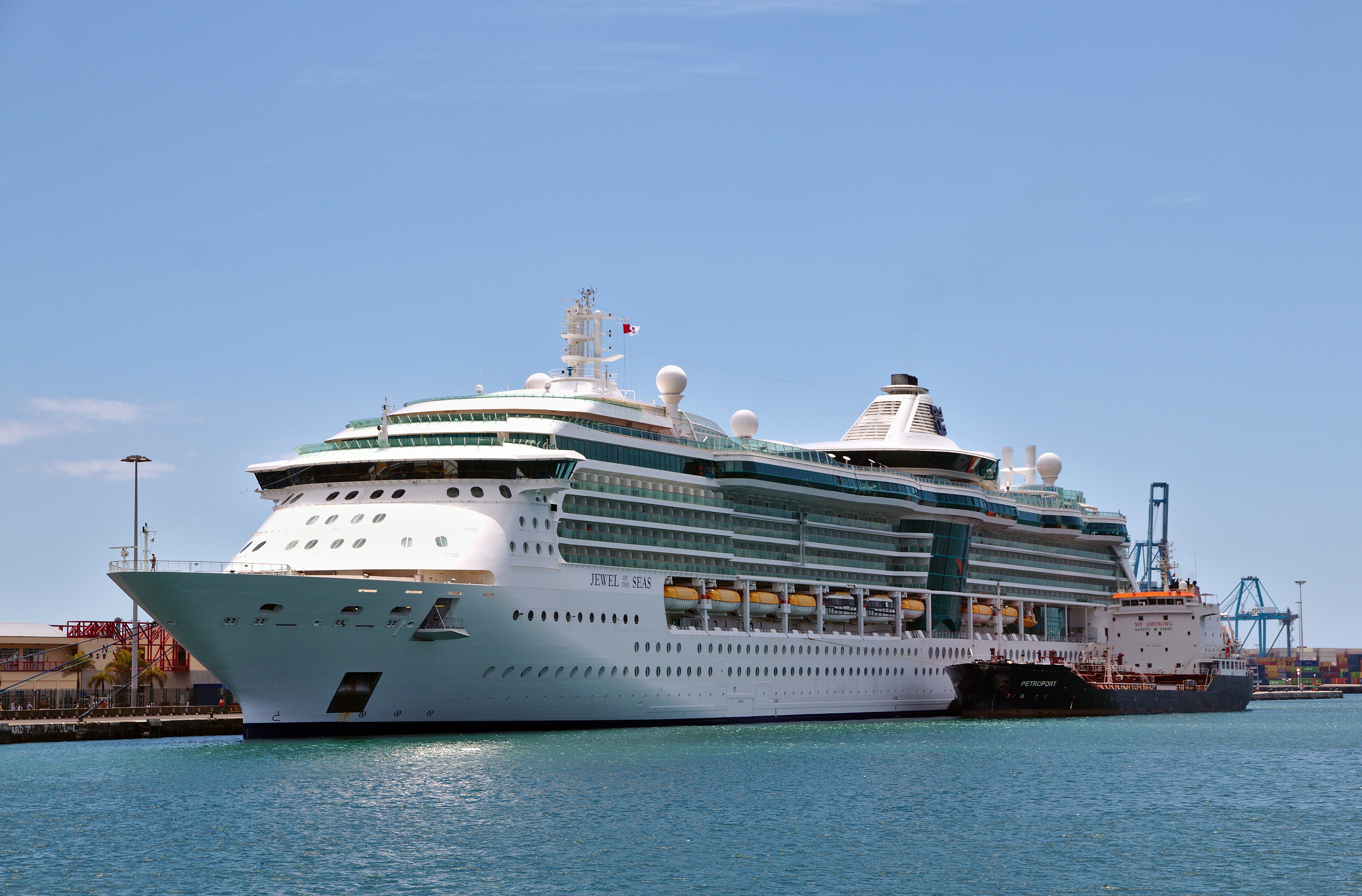 Ein großes weißes Kreuzfahrtschiff namens Jewel of the Seas liegt im Hafen neben einem Tanker.