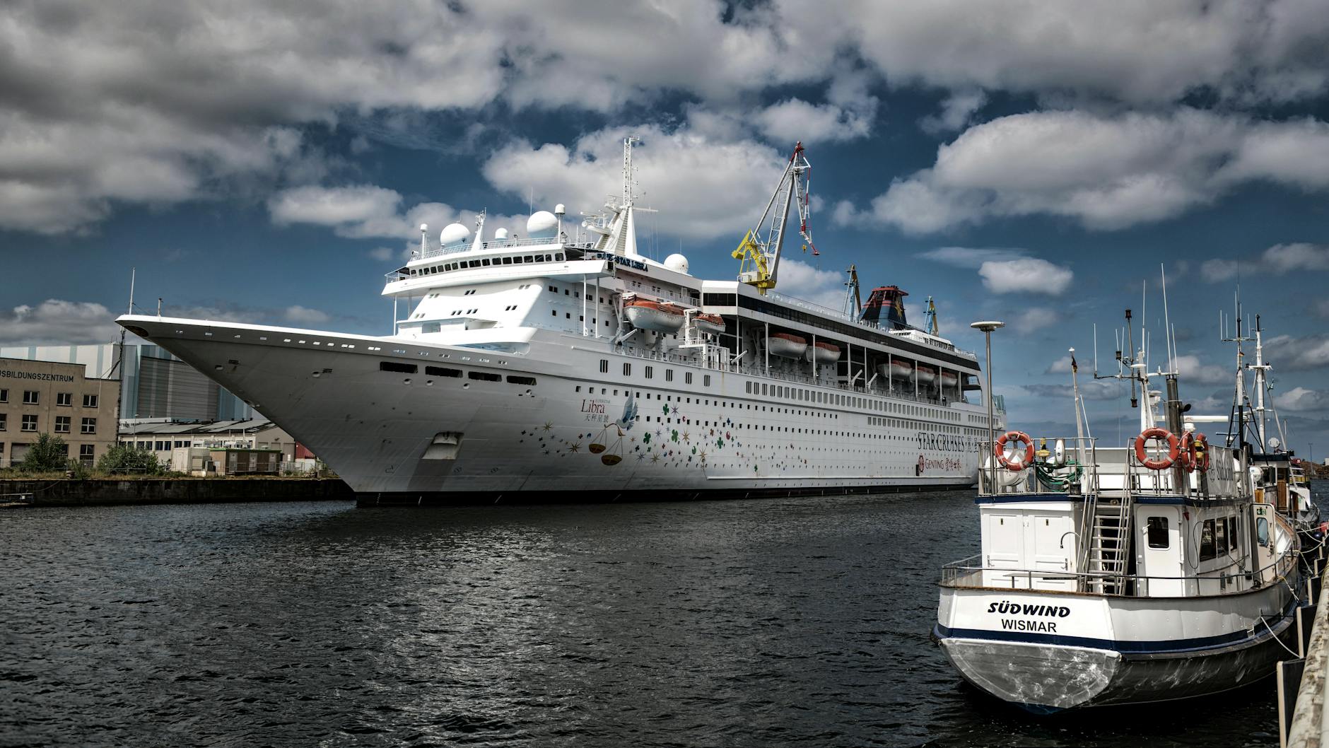 Ein großes weißes Kreuzfahrtschiff liegt im Hafen neben einem kleineren Boot mit dem Namen SÜDWIND WISMAR.