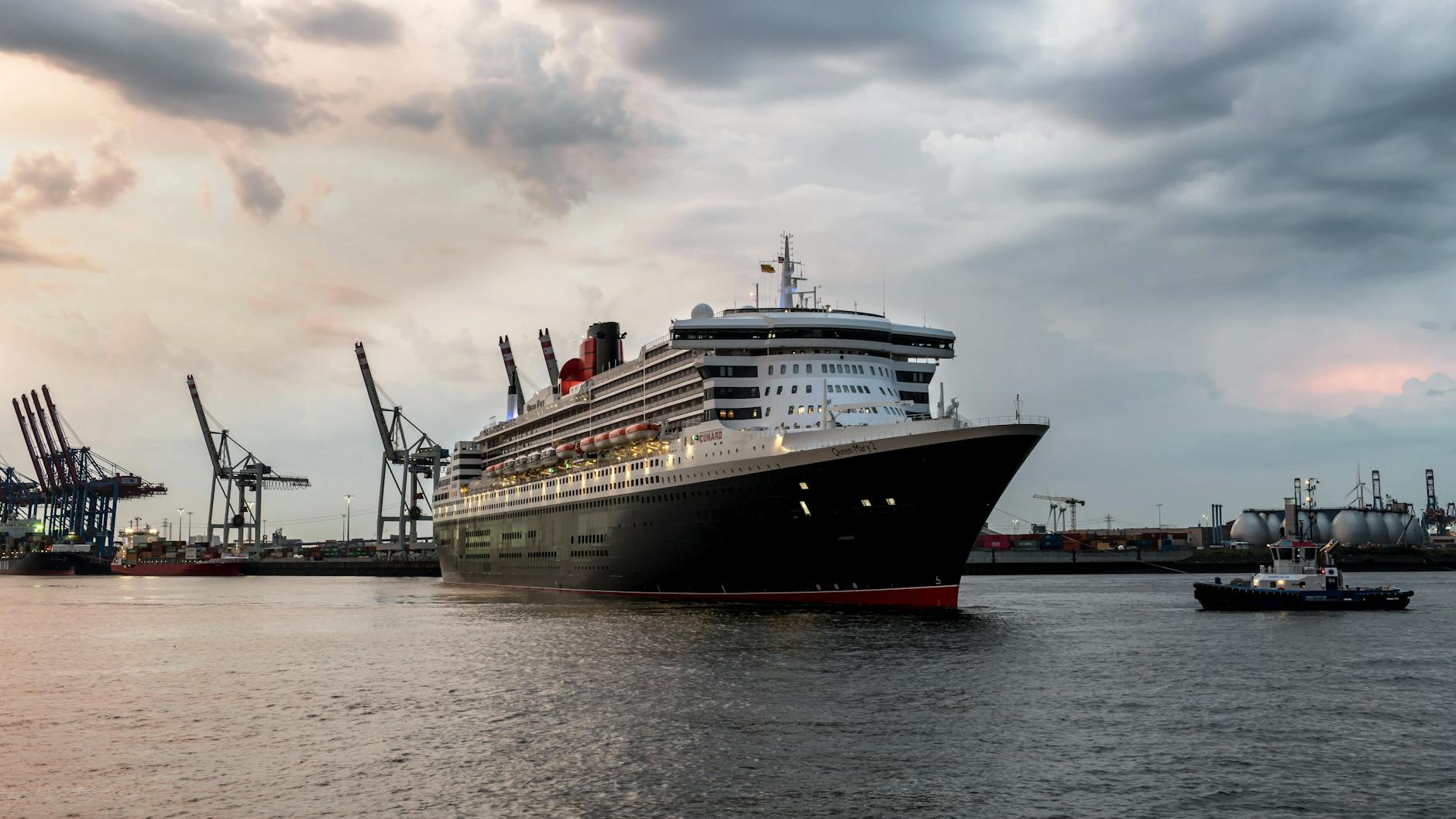 Ein großes Kreuzfahrtschiff mit dem Namen Queen Mary 2 fährt in einem Hafen, begleitet von einem Schlepper.