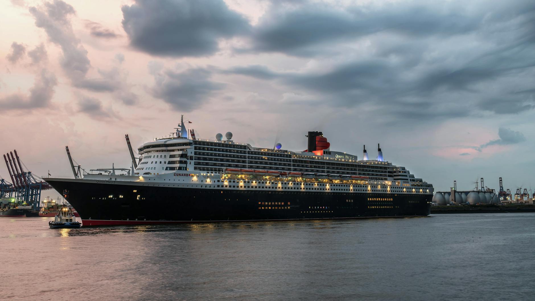 Ein großes Kreuzfahrtschiff mit dem Namen Queen Mary 2 fährt in einem Hafen, im Hintergrund sind Kräne und Industrieanlagen zu sehen.