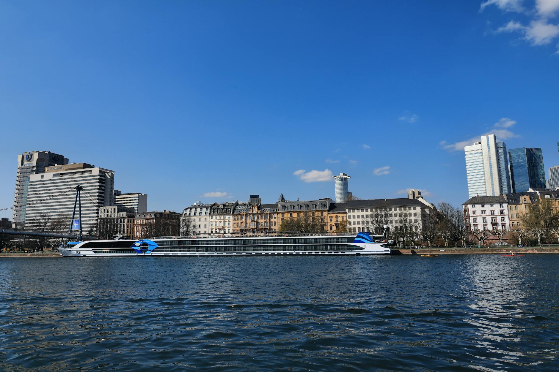 Ein Flusskreuzfahrtschiff liegt am Ufer eines Flusses, im Hintergrund eine Stadtkulisse mit Hochhäusern und historischen Gebäuden.