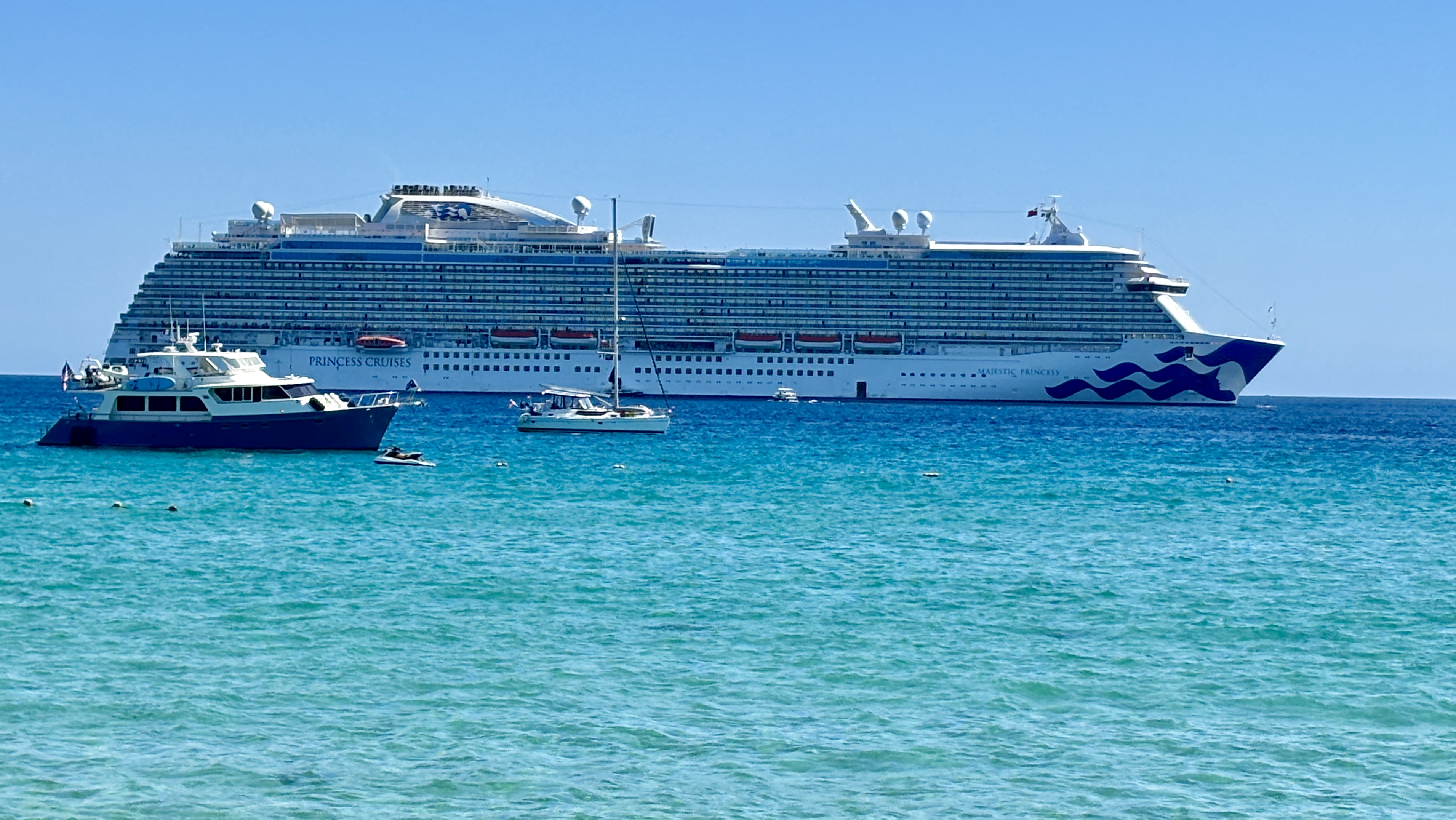 Ein großes Kreuzfahrtschiff der Princess Cruises ankert im türkisfarbenen Meer, daneben liegen eine Yacht und ein Segelboot.