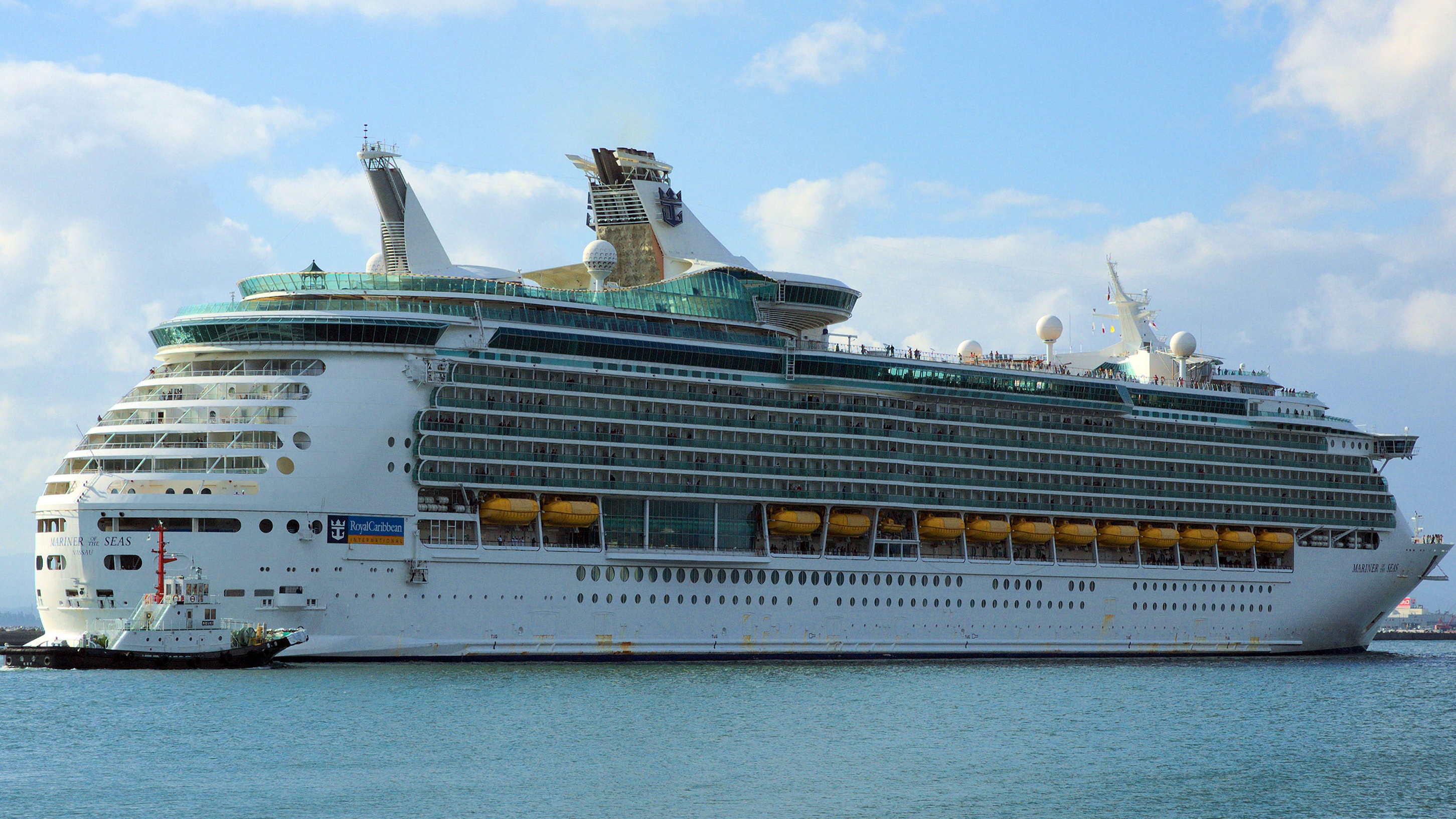 Ein großes Kreuzfahrtschiff namens Mariner of the Seas mit Rettungsbooten an der Seite fährt auf dem Meer, begleitet von einem kleinen Schlepper.