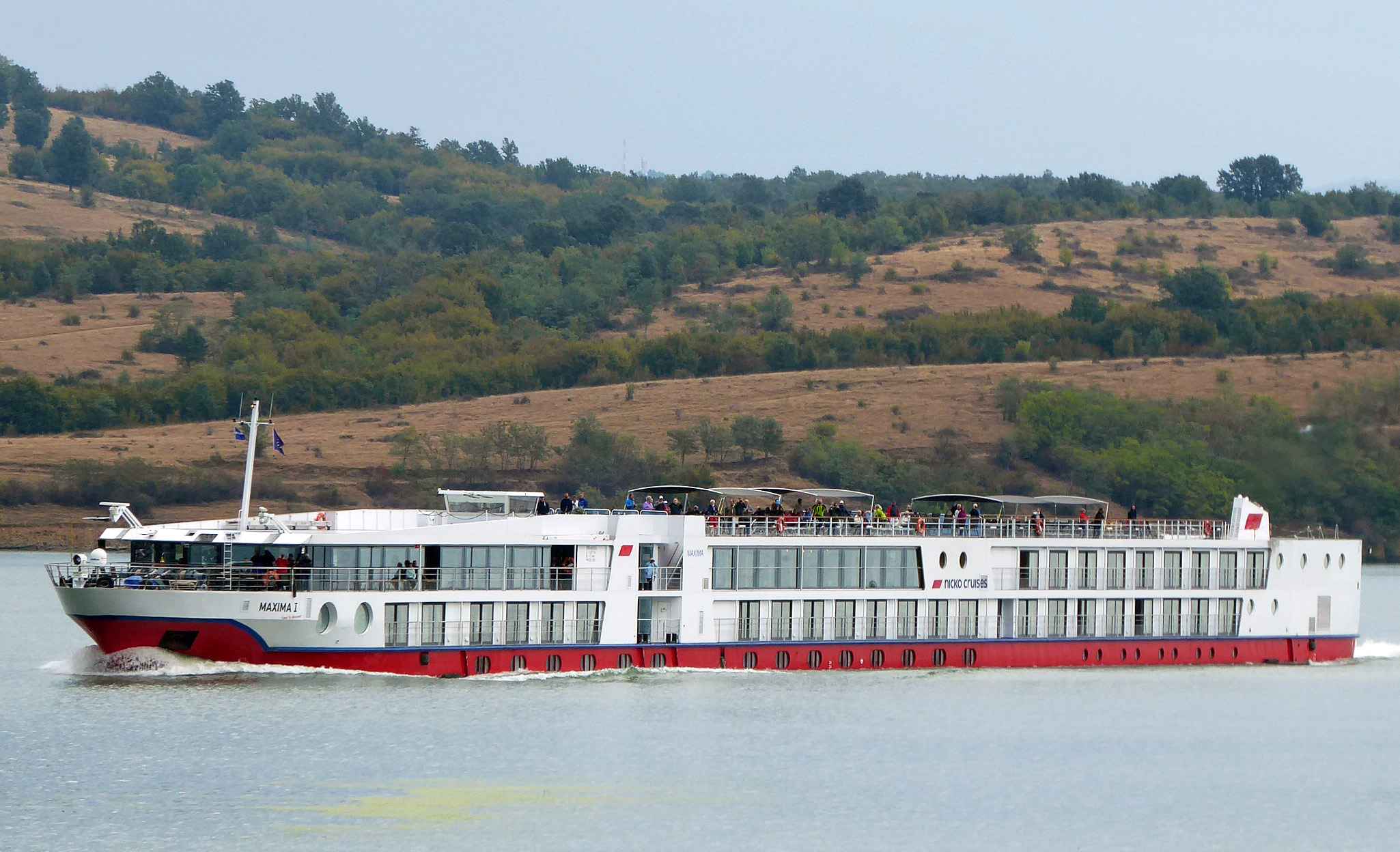 Ein weißes Flusskreuzfahrtschiff mit rotem Rumpf fährt auf einem Fluss, im Hintergrund eine hügelige, bewaldete Landschaft.