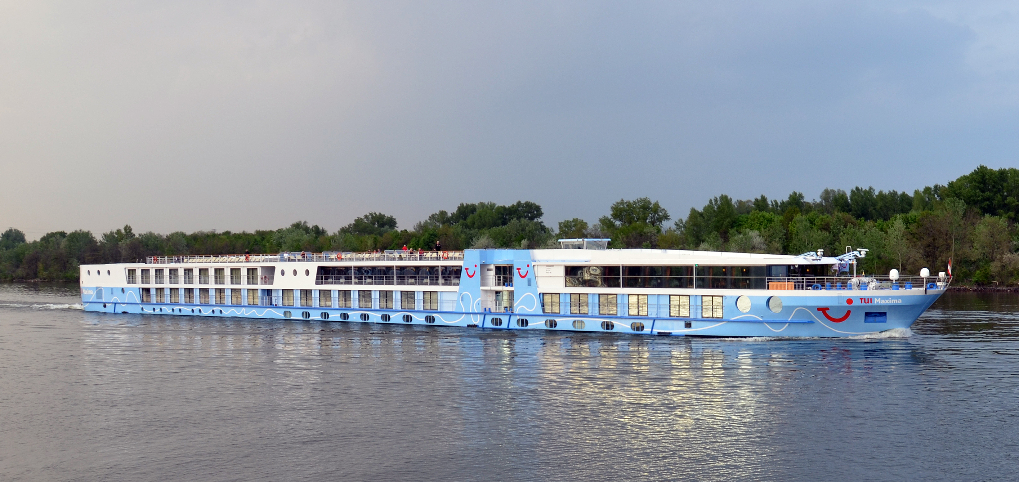 Ein blau-weißes Flusskreuzfahrtschiff namens TUI Maxima fährt auf einem Fluss, mit grünen Bäumen am Ufer im Hintergrund.