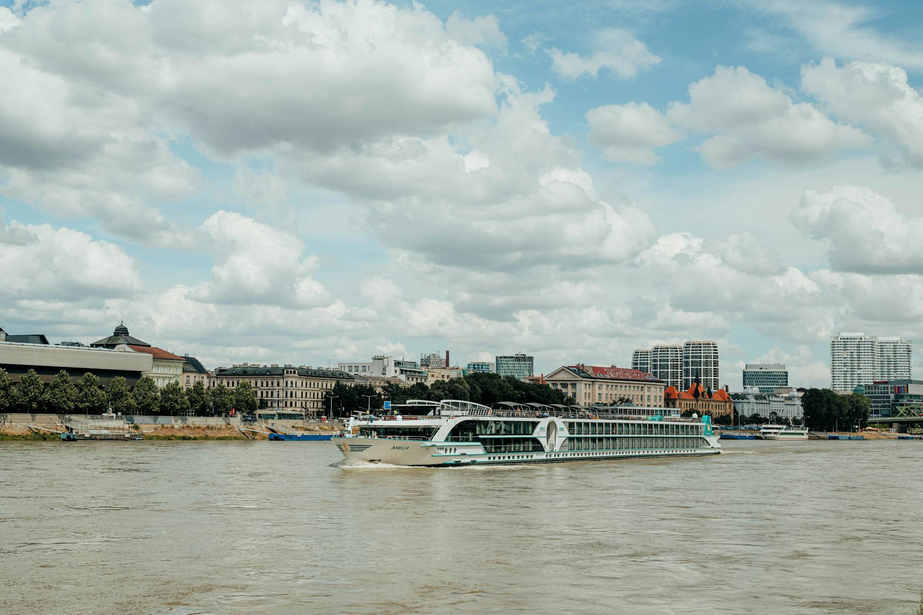 Ein Flusskreuzfahrtschiff fährt auf einem breiten Fluss, im Hintergrund eine Stadtkulisse mit Gebäuden und Bäumen.