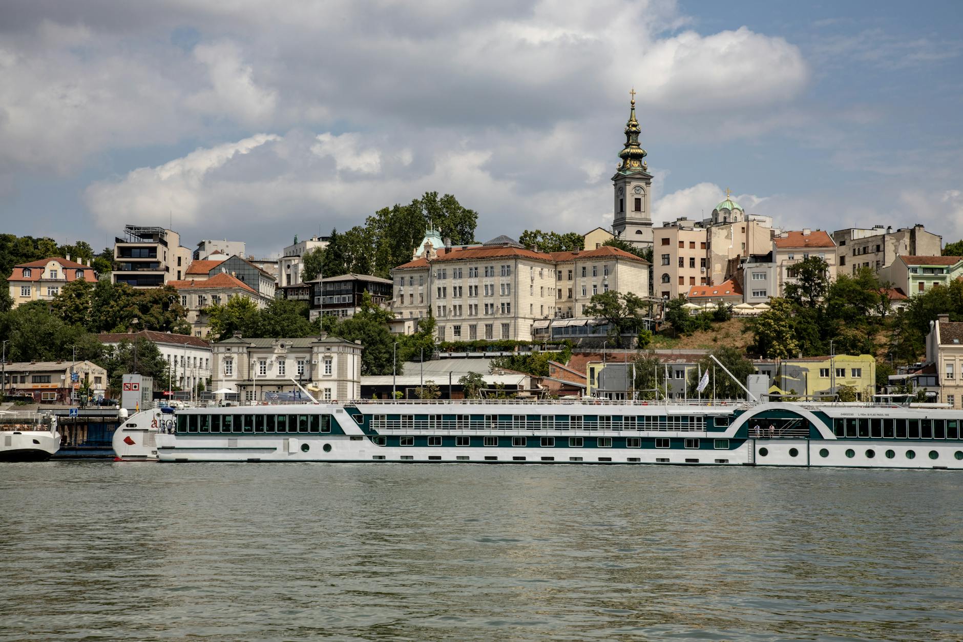 Ein Flusskreuzfahrtschiff liegt im Hafen vor der Kulisse der Stadt Belgrad mit ihren Gebäuden und einem Kirchturm.