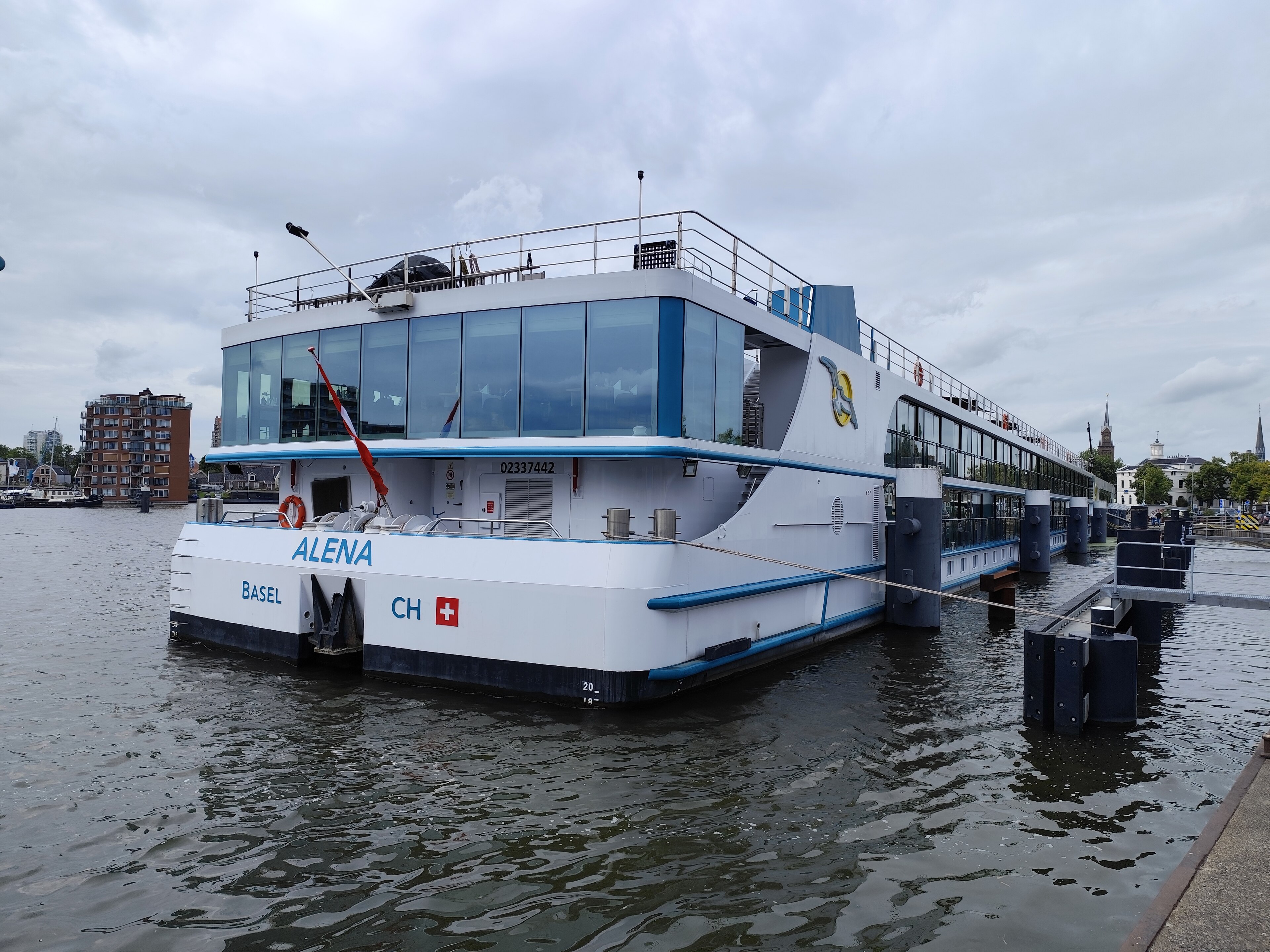 Ein weißes Flusskreuzfahrtschiff mit dem Namen ALENA und der Aufschrift BASEL sowie der Schweizer Flagge liegt im Hafen an einer Anlegestelle.