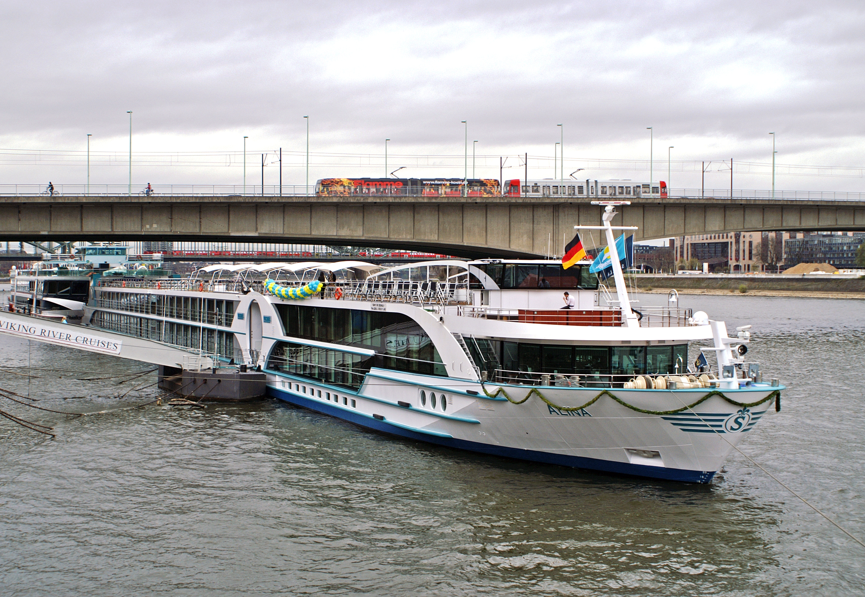Ein Flusskreuzfahrtschiff namens Alina liegt im Wasser, während im Hintergrund eine Brücke mit Straßenbahn und Radfahrern zu sehen ist.