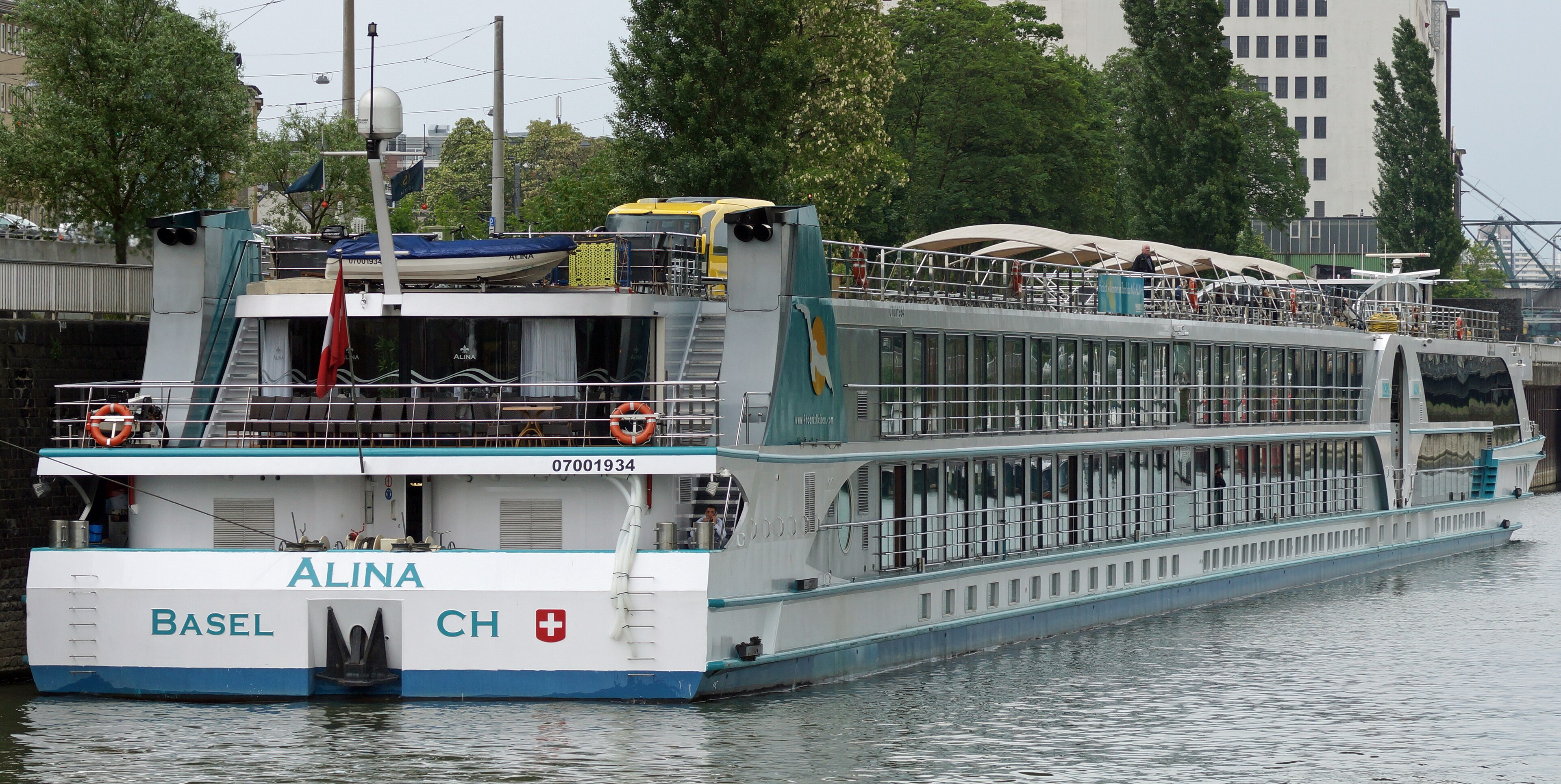 Ein Flusskreuzfahrtschiff mit dem Namen ALINA und der Kennung BASEL CH liegt im Wasser vor einer Stadtkulisse.