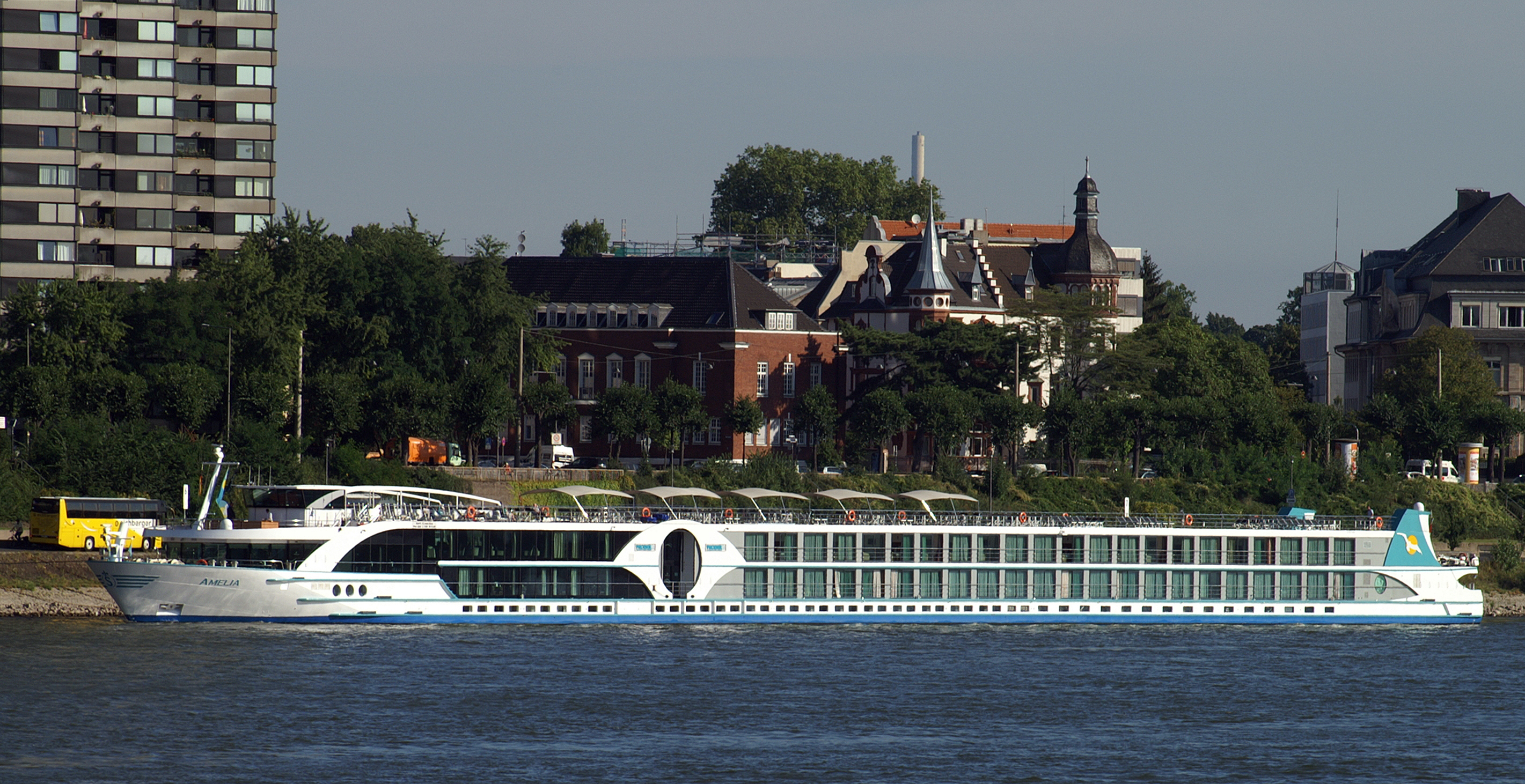 Ein weisses Flusskreuzfahrtschiff mit dem Namen AMELIA faehrt auf einem Fluss, im Hintergrund sind Gebaeude und Baeume zu sehen.
