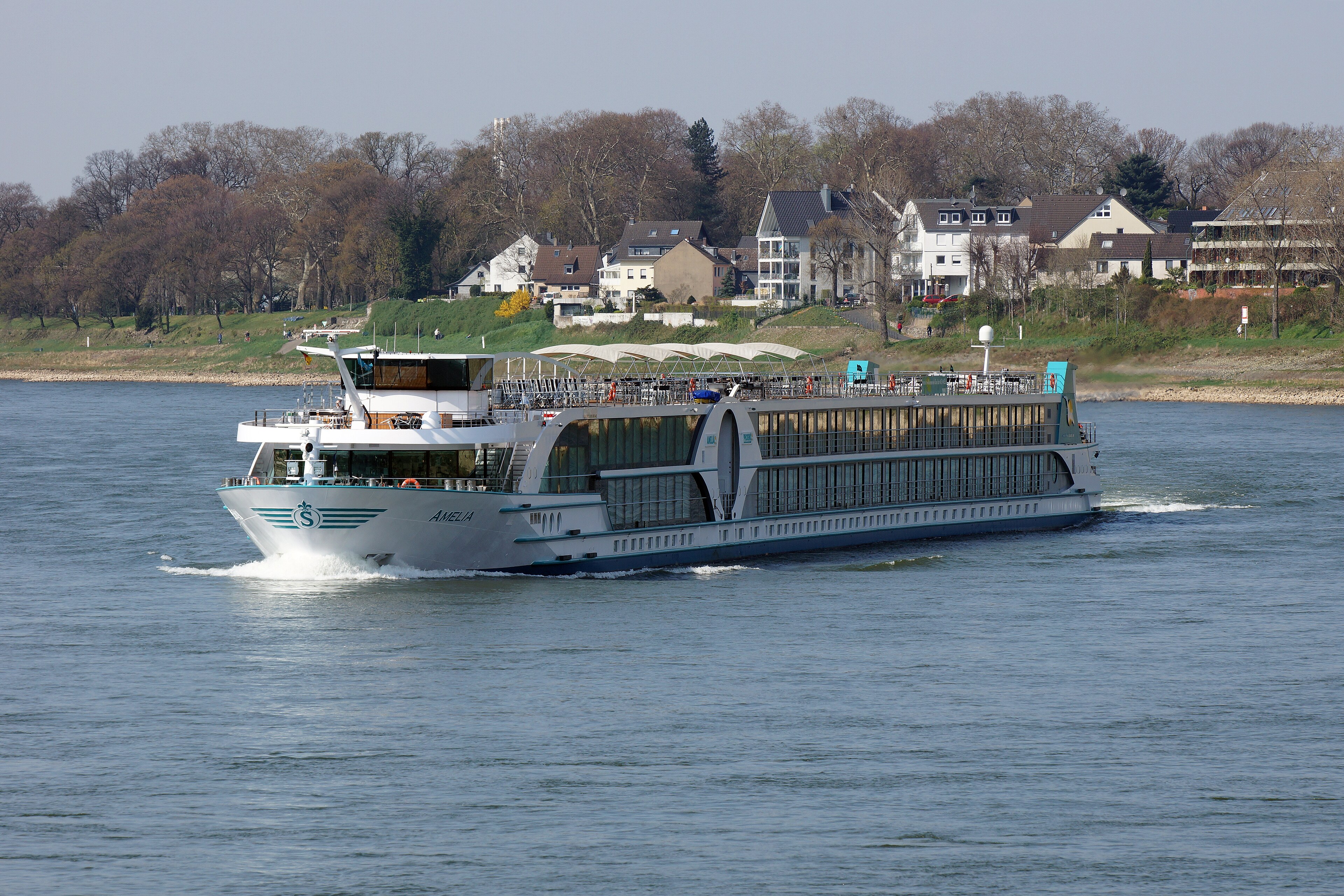 Ein Flusskreuzfahrtschiff mit dem Namen AMELIA fährt auf einem Fluss, im Hintergrund sind Häuser und Bäume zu sehen.