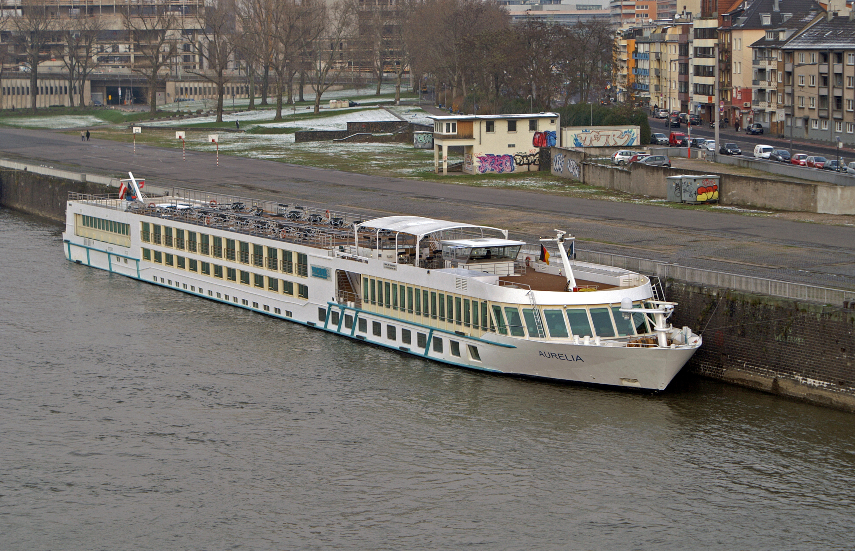 Ein weisses Flusskreuzfahrtschiff mit dem Namen Aurelia liegt an einer Backsteinmauer in einem Hafen, im Hintergrund sind Gebaeude und Baeume zu sehen.