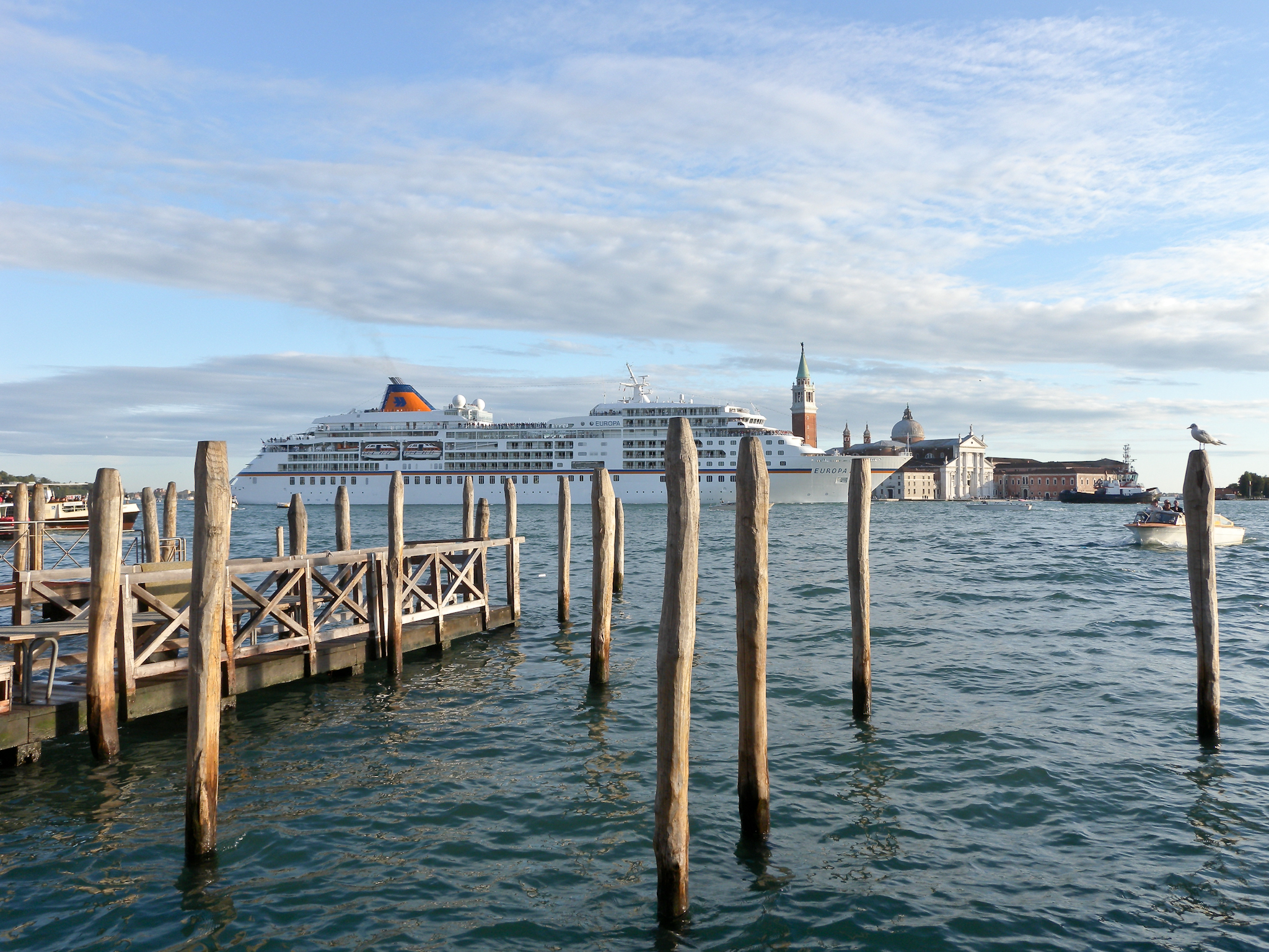 Ein Kreuzfahrtschiff liegt im Wasser vor einer Stadtkulisse mit Kirchturm und Kuppel, im Vordergrund sind Holzpfähle und ein Steg zu sehen.