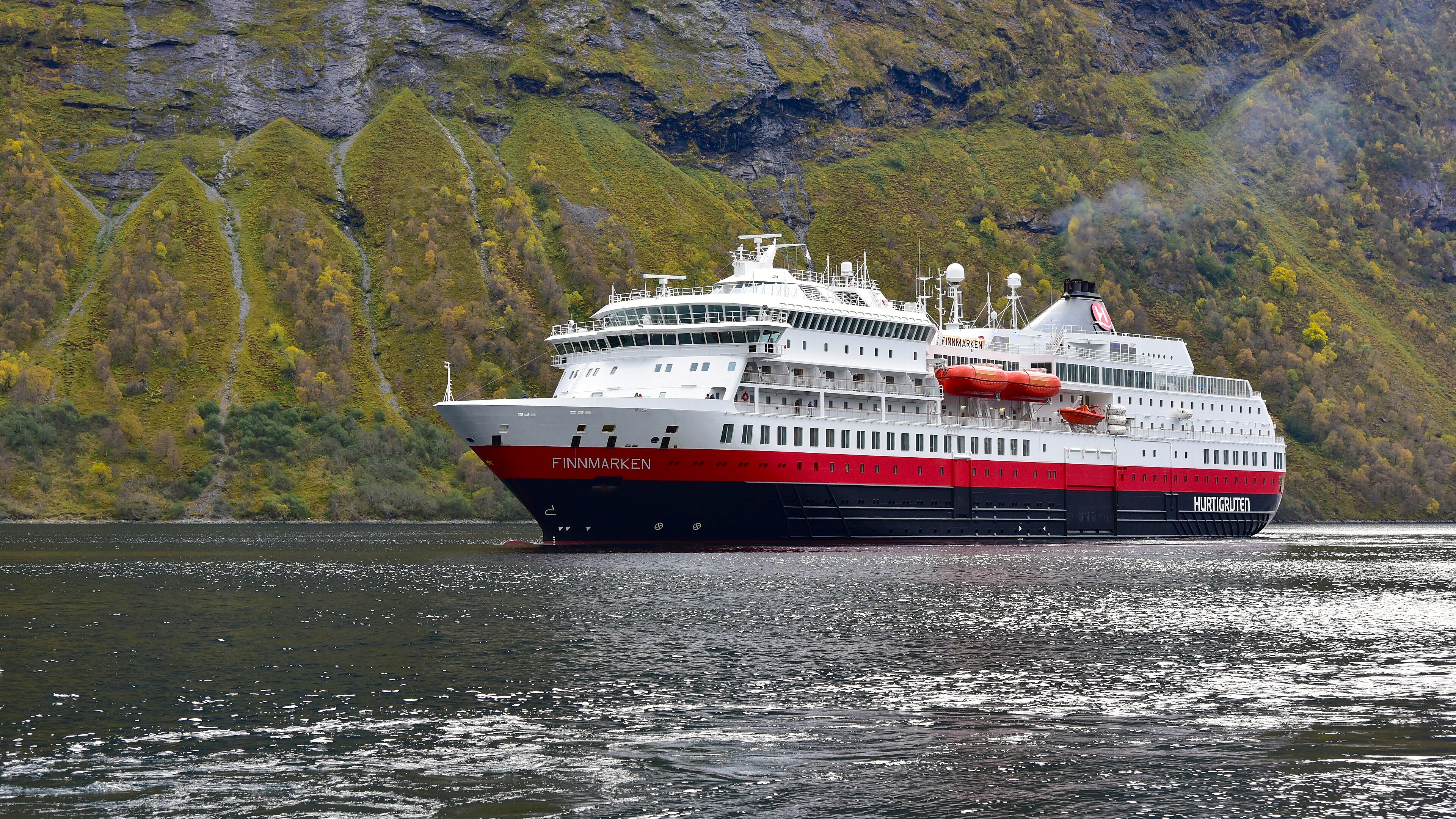 Ein Hurtigruten-Passagierschiff namens Finnmarken fährt auf einem Fjord vor einer bewaldeten Bergkulisse.