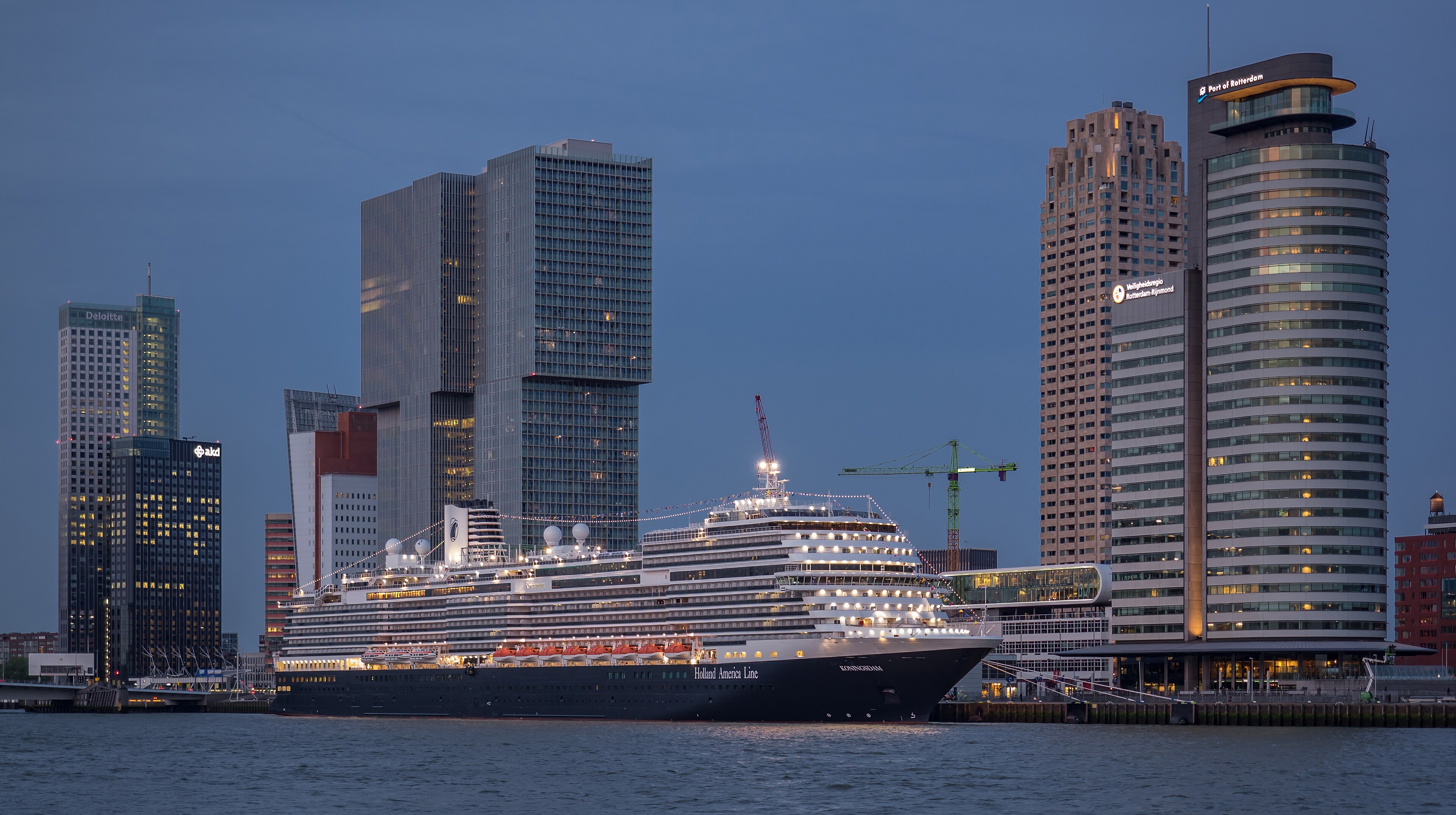 Ein großes Kreuzfahrtschiff der Holland America Line mit dem Namen Koningsdam liegt im Hafen von Rotterdam vor einer Skyline mit modernen Hochhäusern.