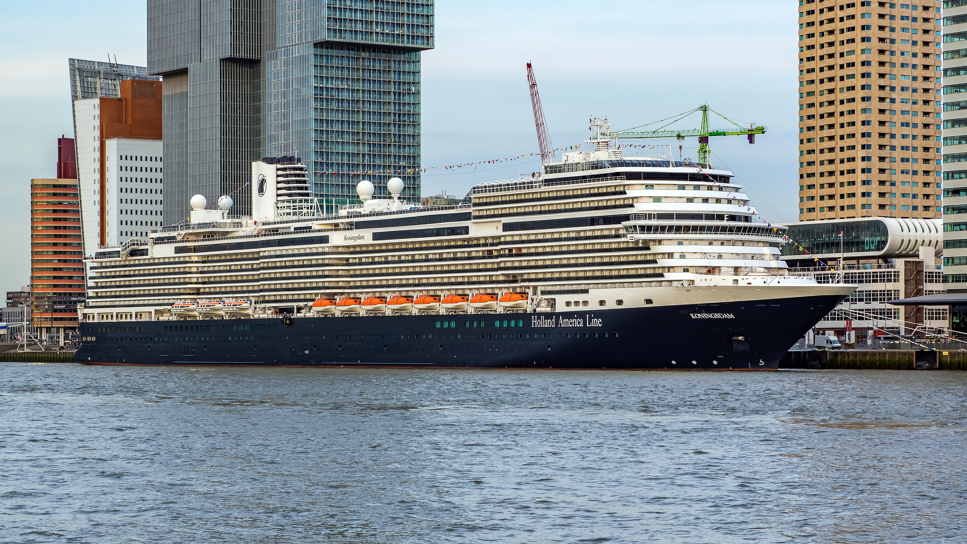 Ein großes Kreuzfahrtschiff der Holland America Line mit dem Namen Koningsdam liegt im Hafen vor einer modernen Stadtkulisse mit hohen Gebäuden und Kränen.