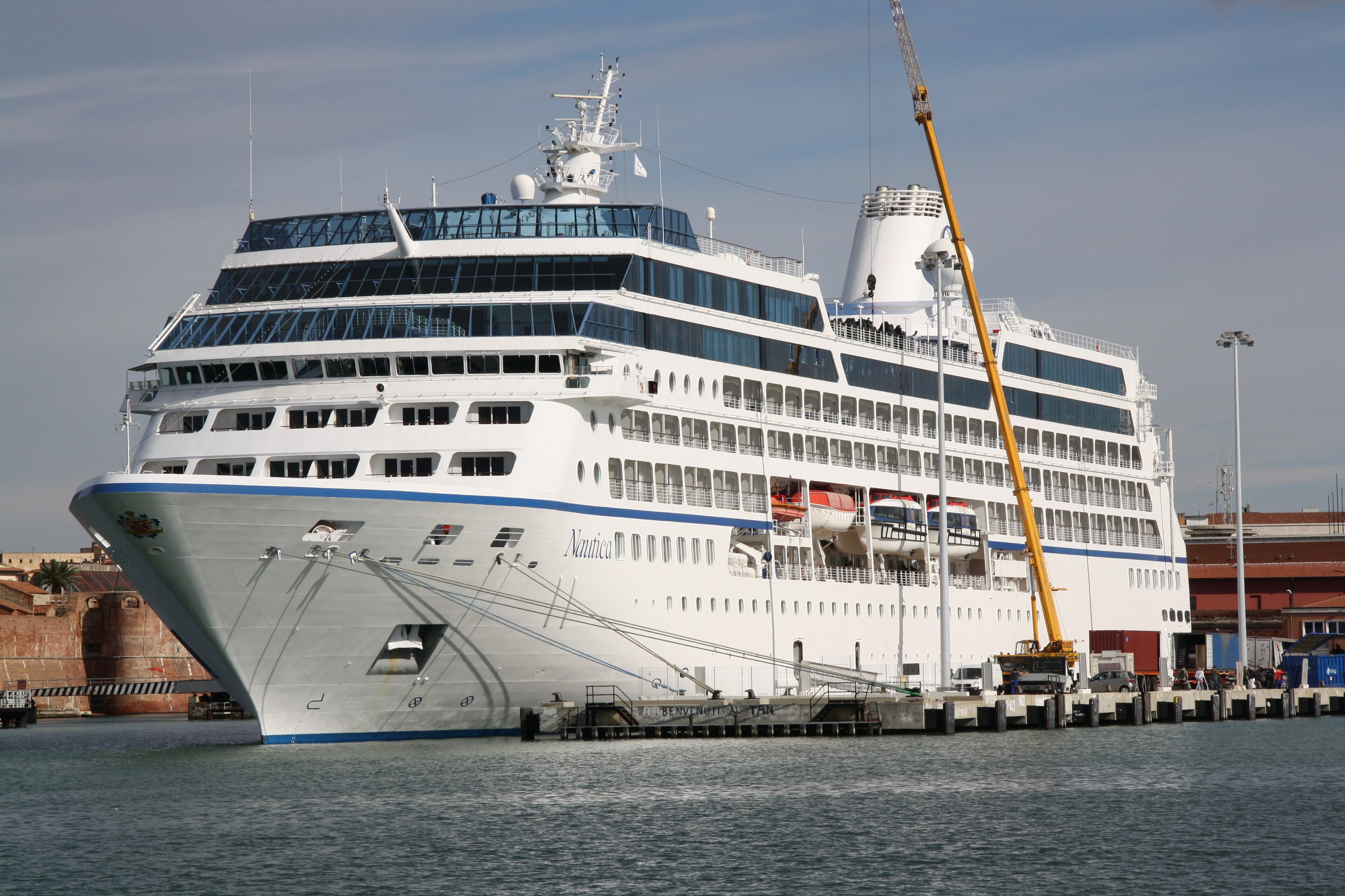 Ein großes weißes Kreuzfahrtschiff liegt im Hafen an einem Pier, daneben steht ein gelber Kran.