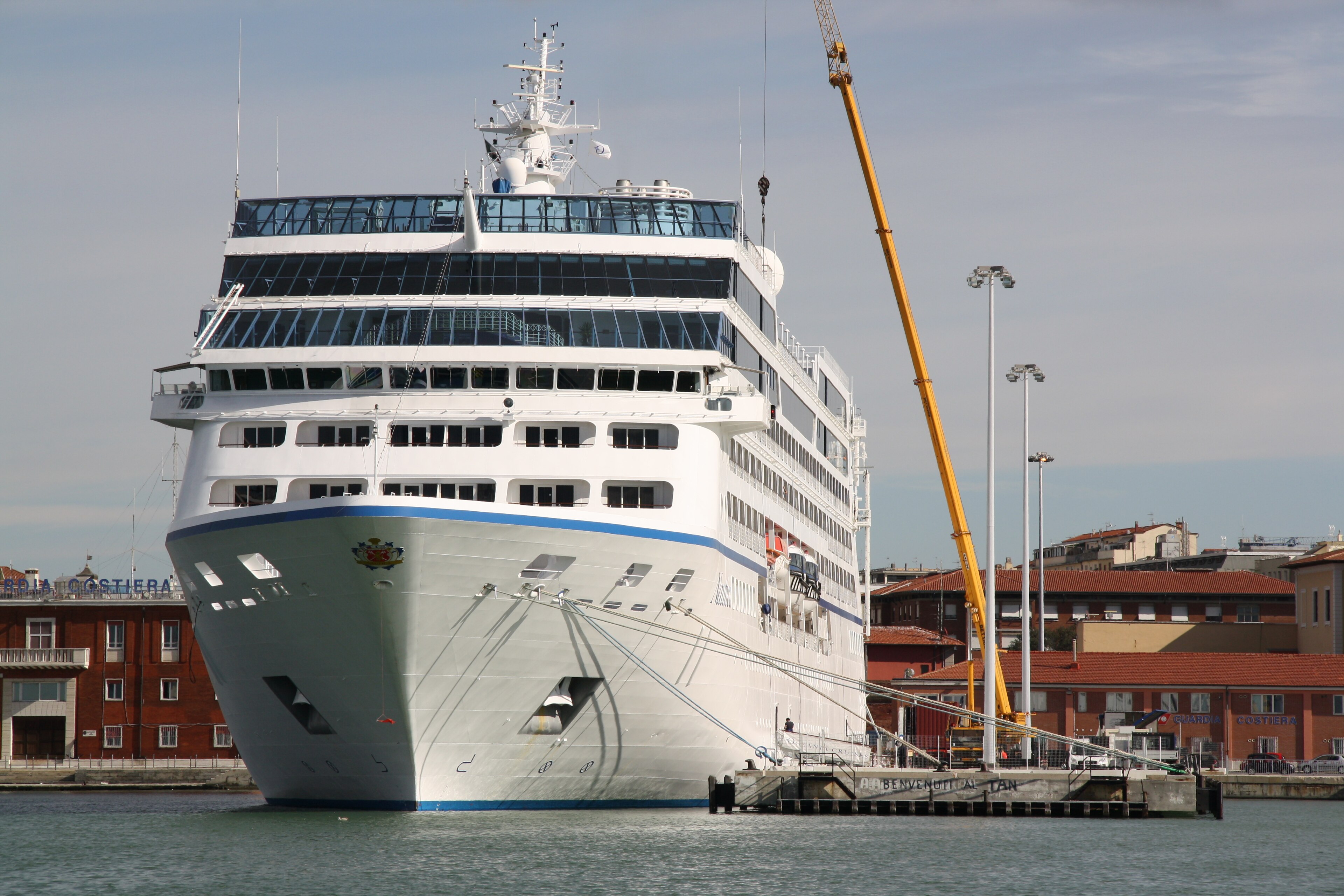 Ein großes weißes Kreuzfahrtschiff liegt im Hafen an einem Pier, daneben steht ein gelber Kran.
