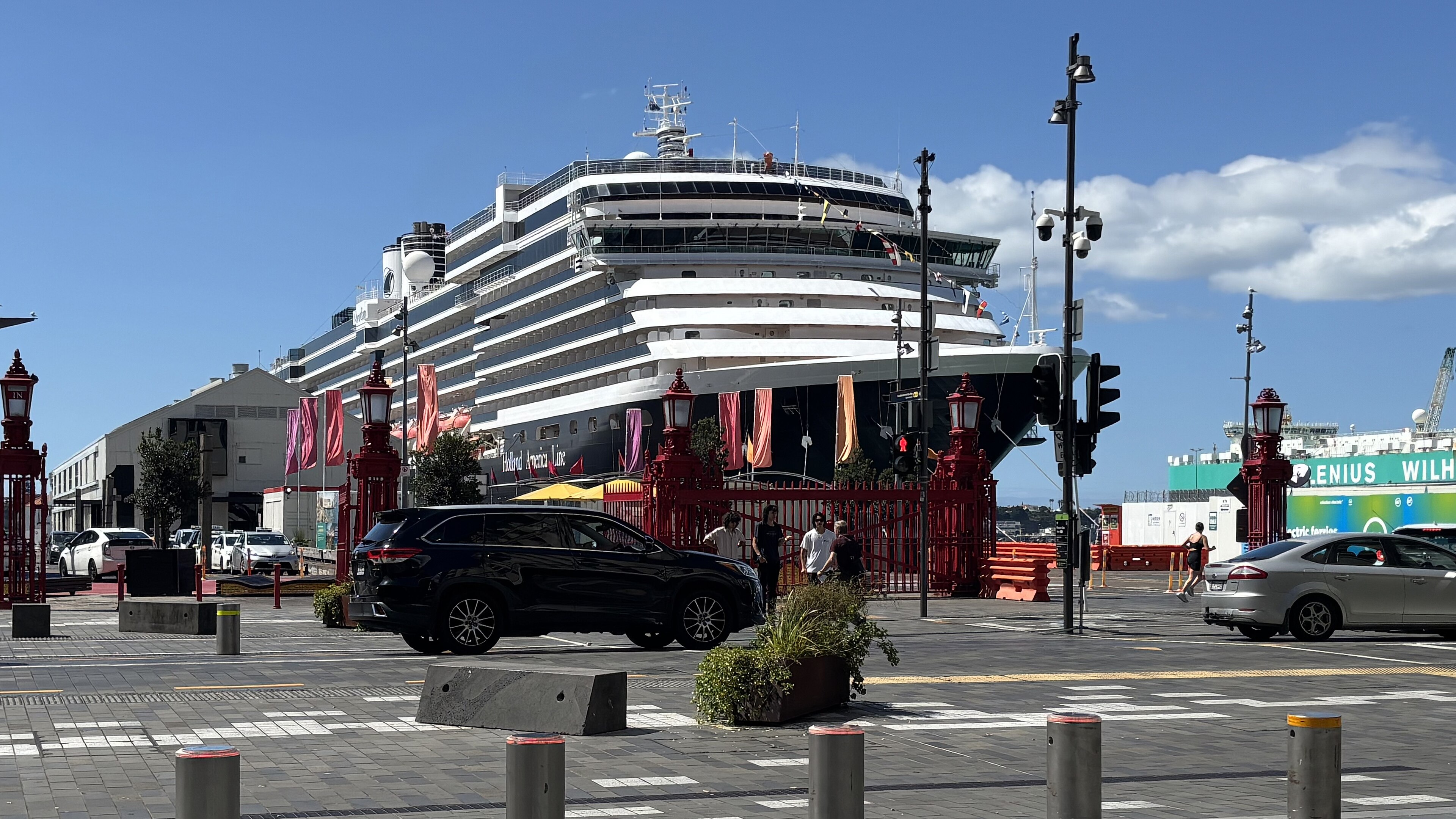 Ein großes Kreuzfahrtschiff der Holland America Line liegt im Hafen, daneben stehen Autos und Menschen.
