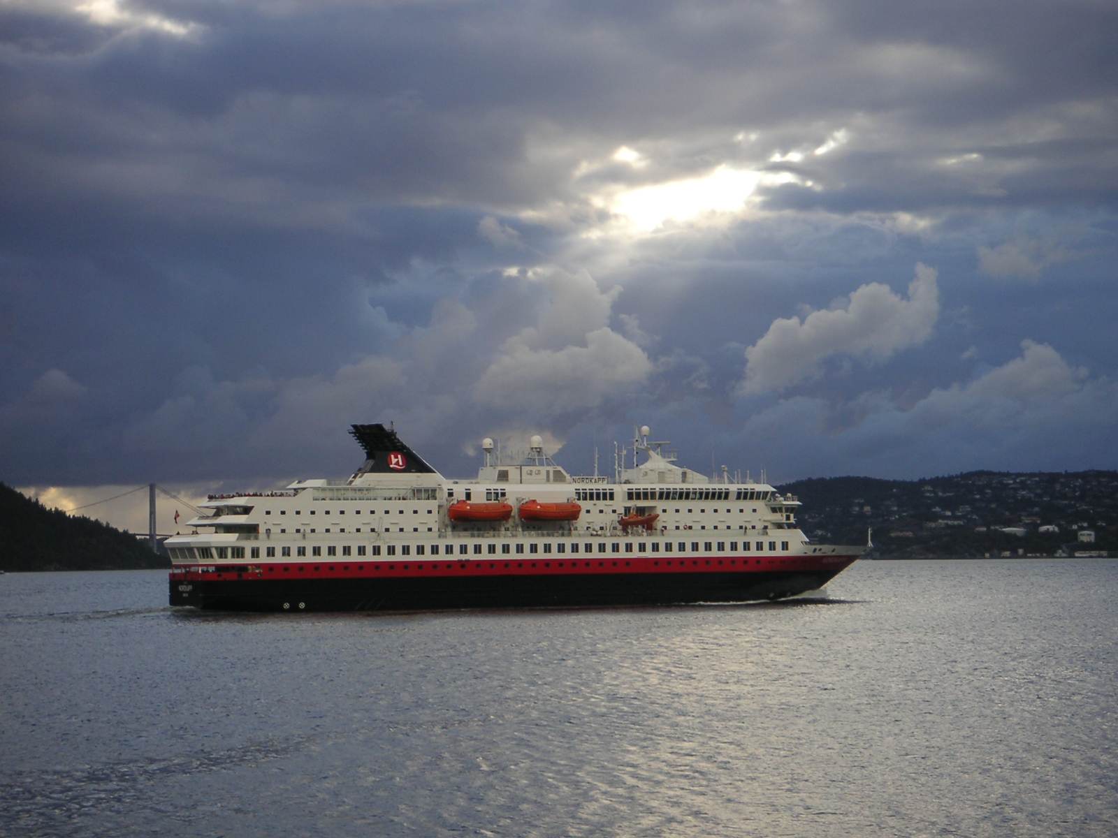 Ein weisses Kreuzfahrtschiff mit rotem Rumpf faehrt auf dem Meer unter einem dramatischen Himmel.
