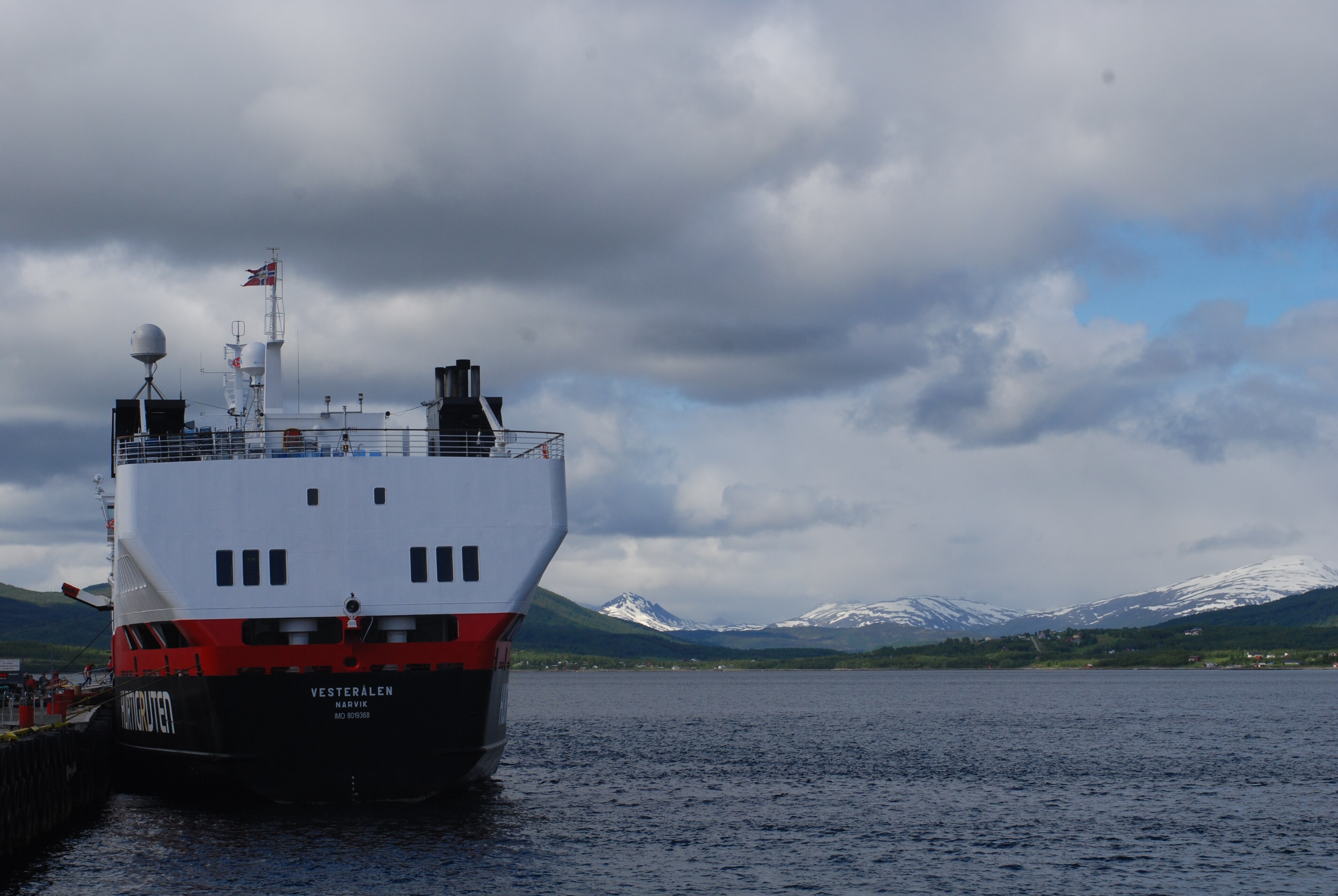 Ein großes weißes und rotes Schiff liegt im Hafen vor schneebedeckten Bergen und einem ruhigen Meer.