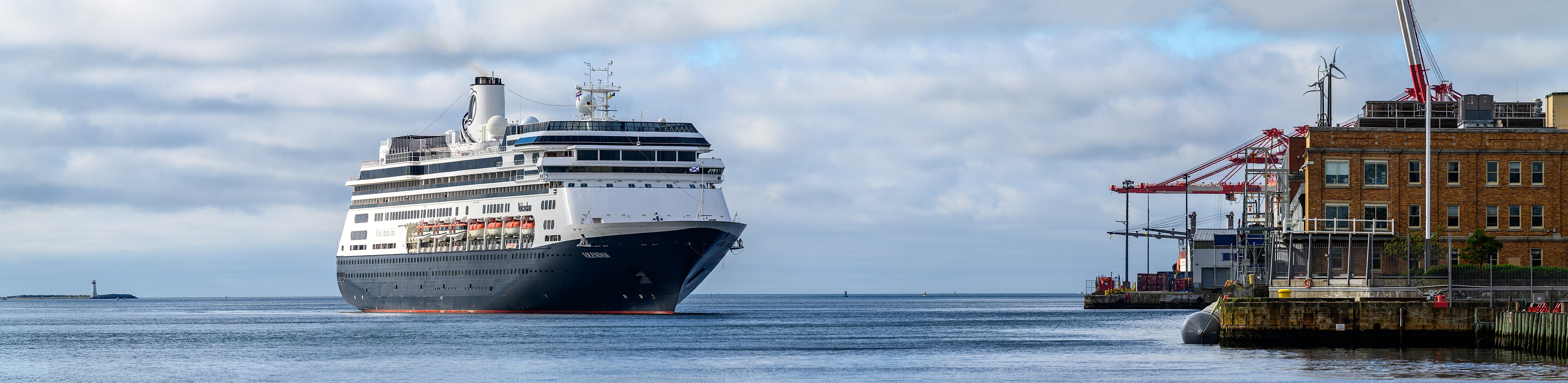 Ein großes Kreuzfahrtschiff fährt in einen Hafen ein, im Hintergrund sind Kräne und ein Leuchtturm zu sehen.