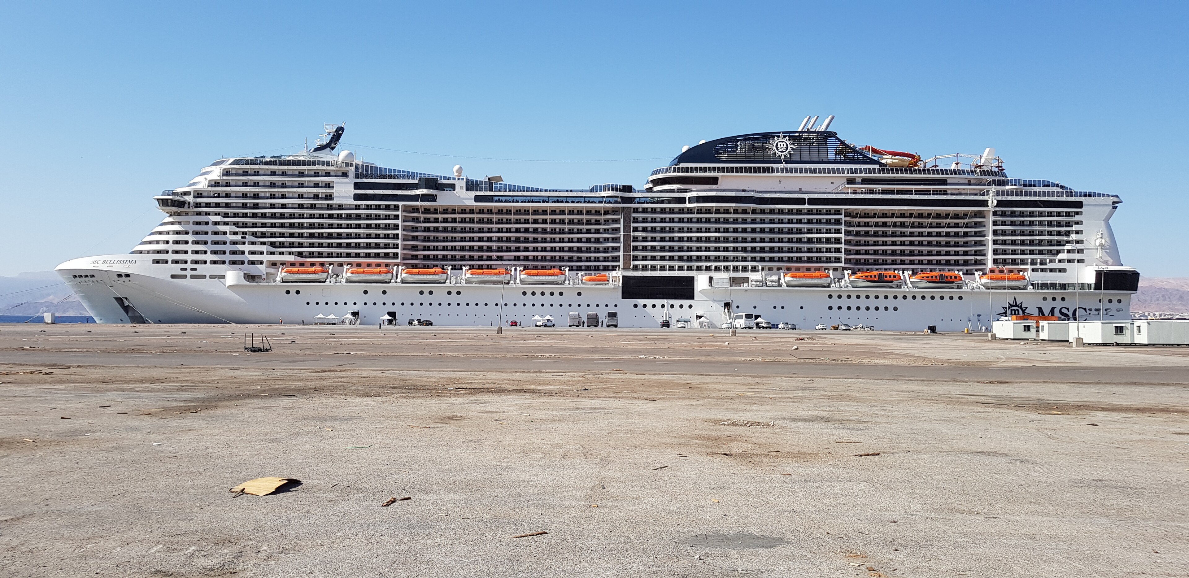 Ein großes weißes Kreuzfahrtschiff liegt im Hafen vor einer staubigen Uferpromenade.