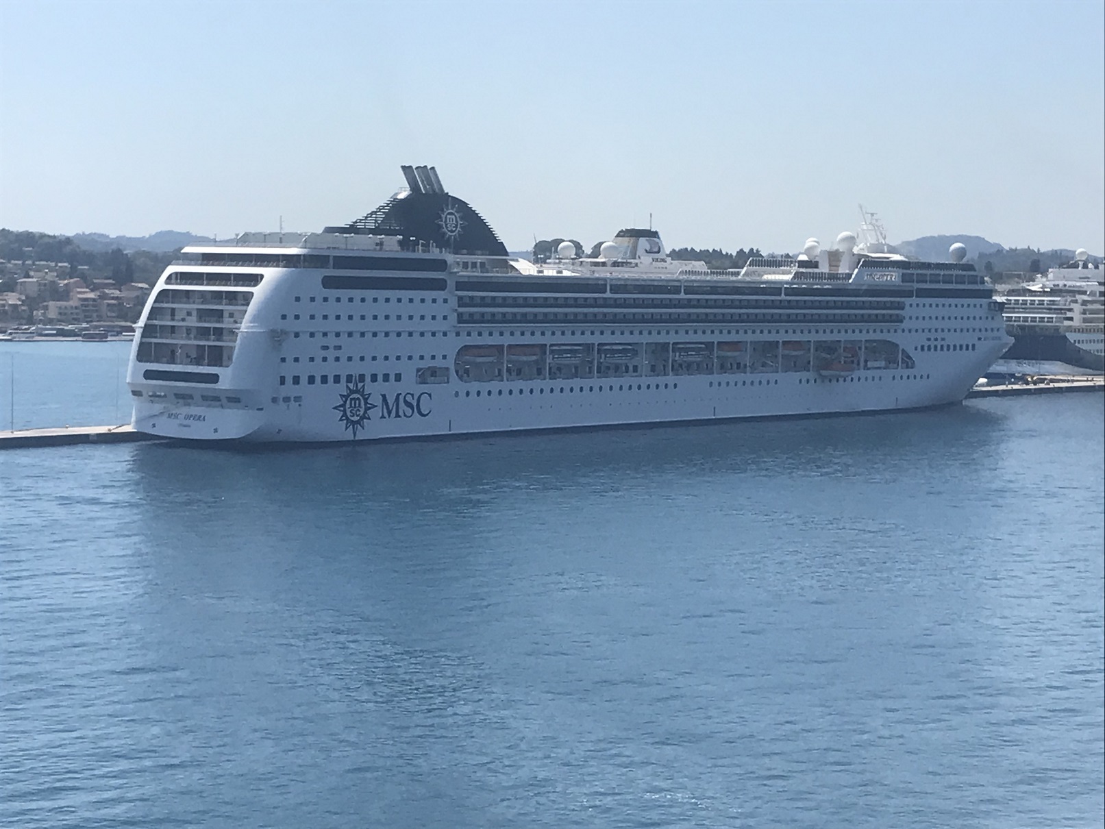 Ein großes weißes Kreuzfahrtschiff mit dem Namen MSC Opera liegt an einem Kai im blauen Wasser.