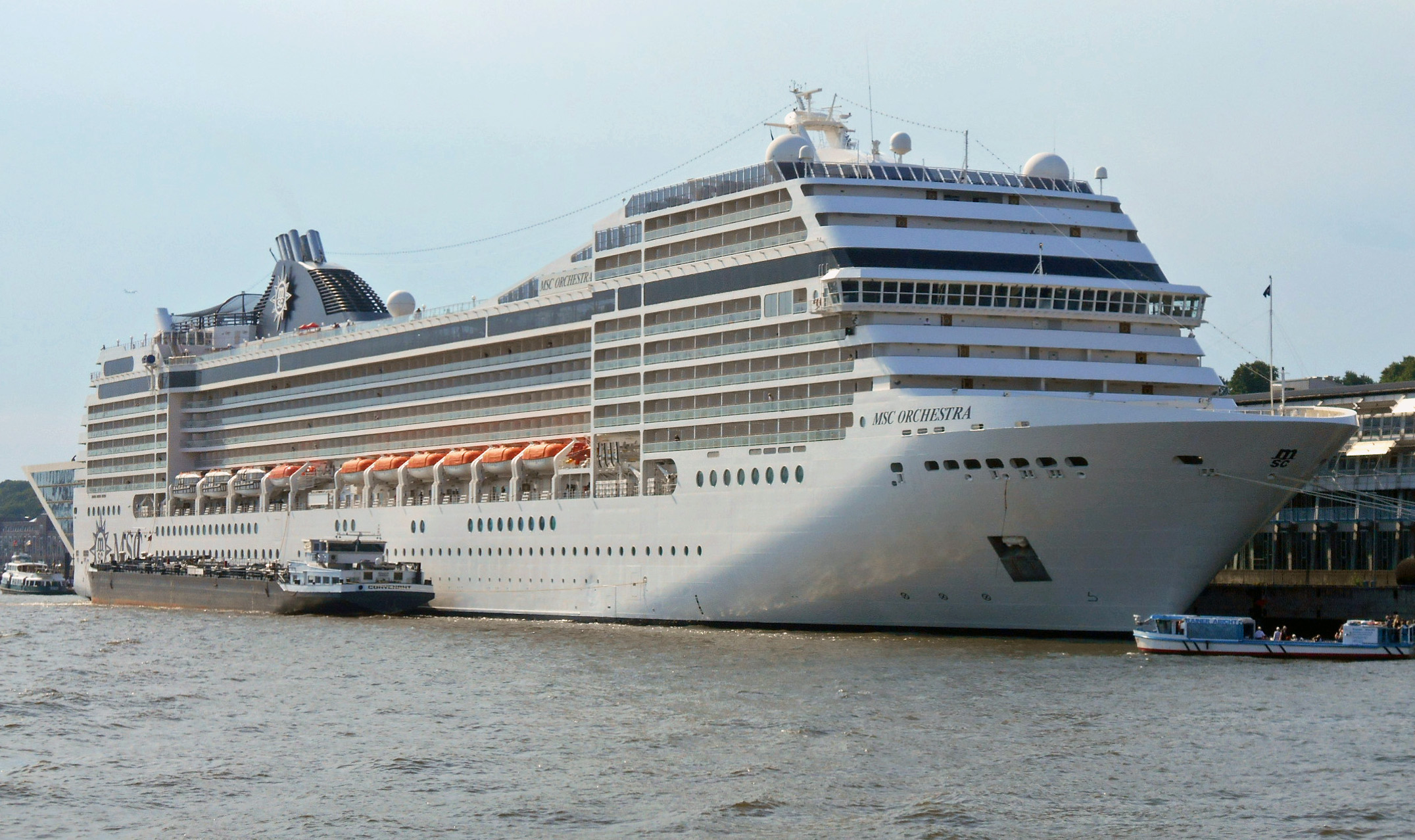 Ein großes weißes Kreuzfahrtschiff namens MSC Orchestra liegt im Hafen neben einem Frachtschiff.