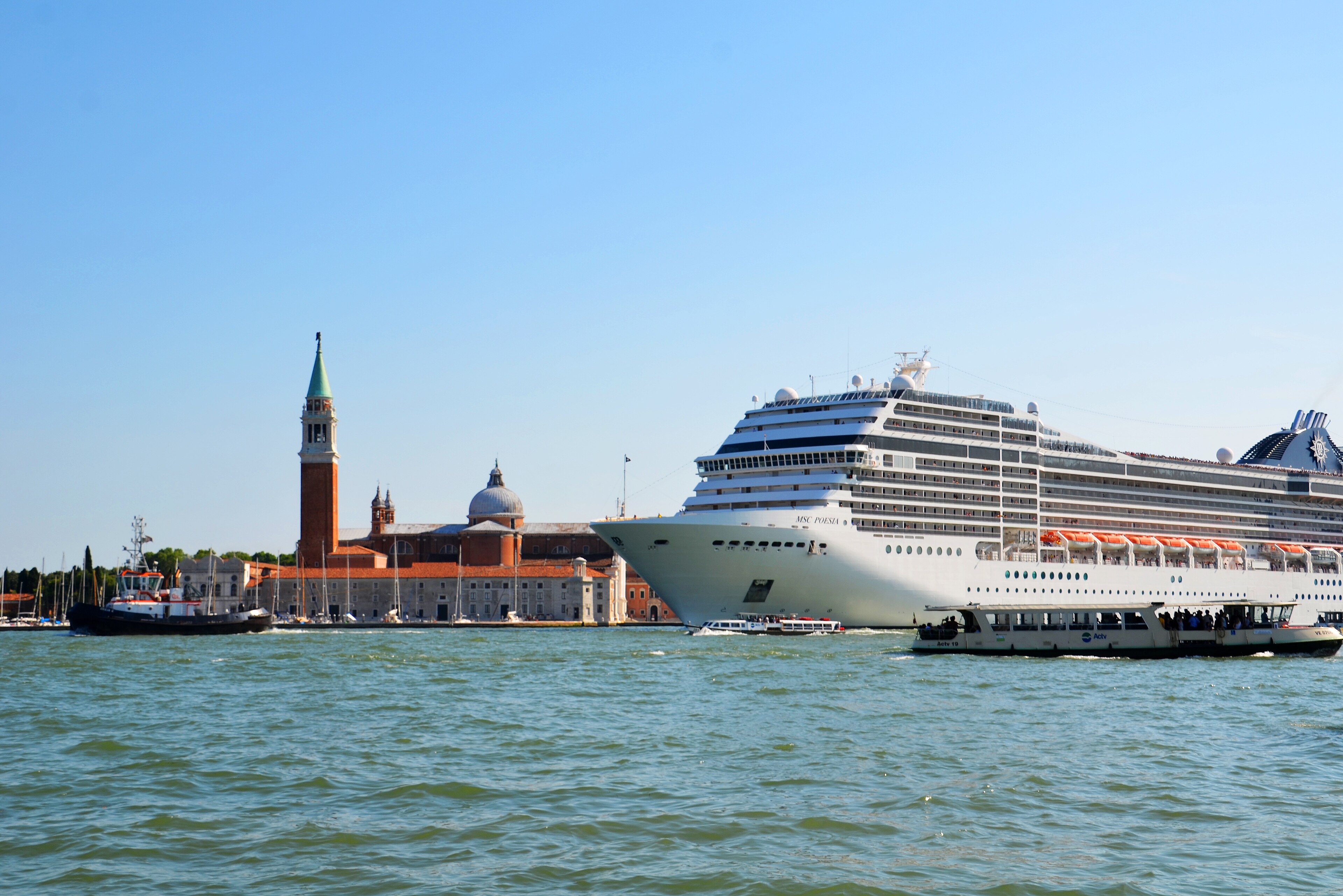 Ein grosses Kreuzfahrtschiff faehrt in Venedig auf dem Wasser, im Hintergrund sind Gebaeude und ein Kirchturm zu sehen.