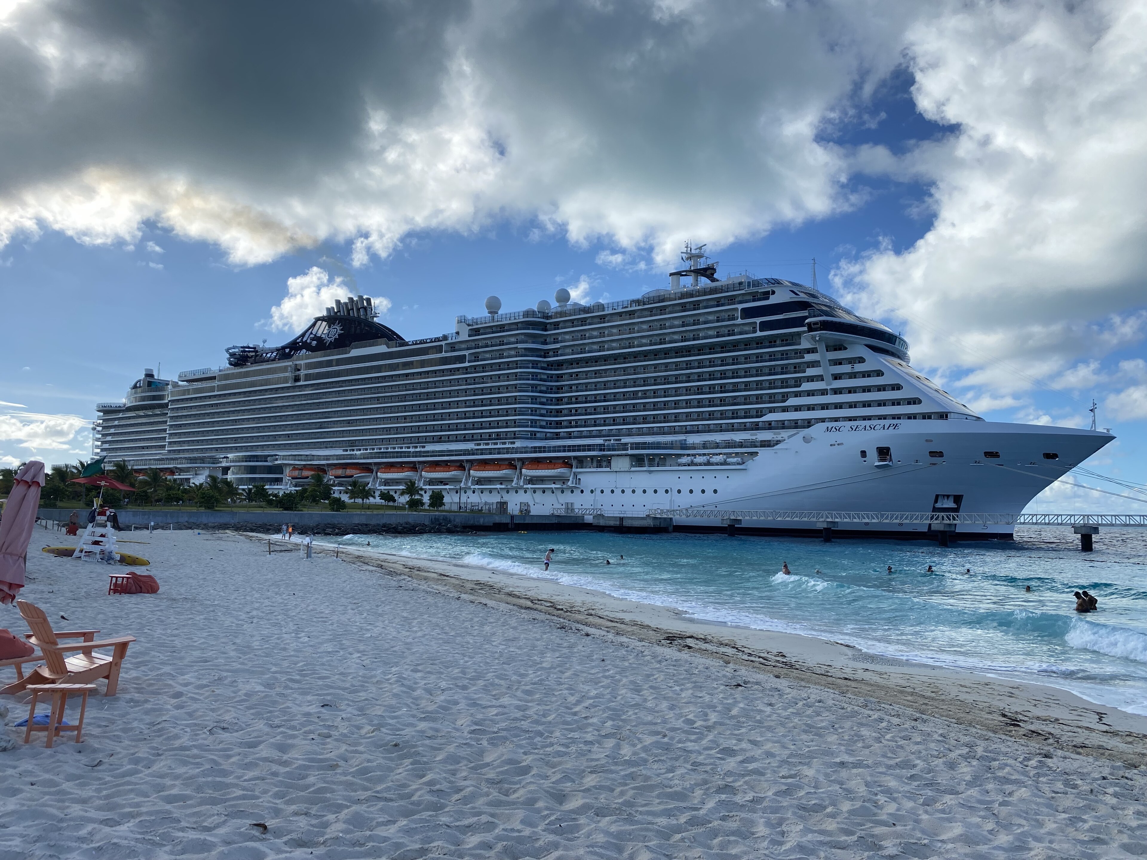 Ein großes Kreuzfahrtschiff liegt im Hafen neben einem Sandstrand mit türkisfarbenem Wasser und einigen Badegästen.