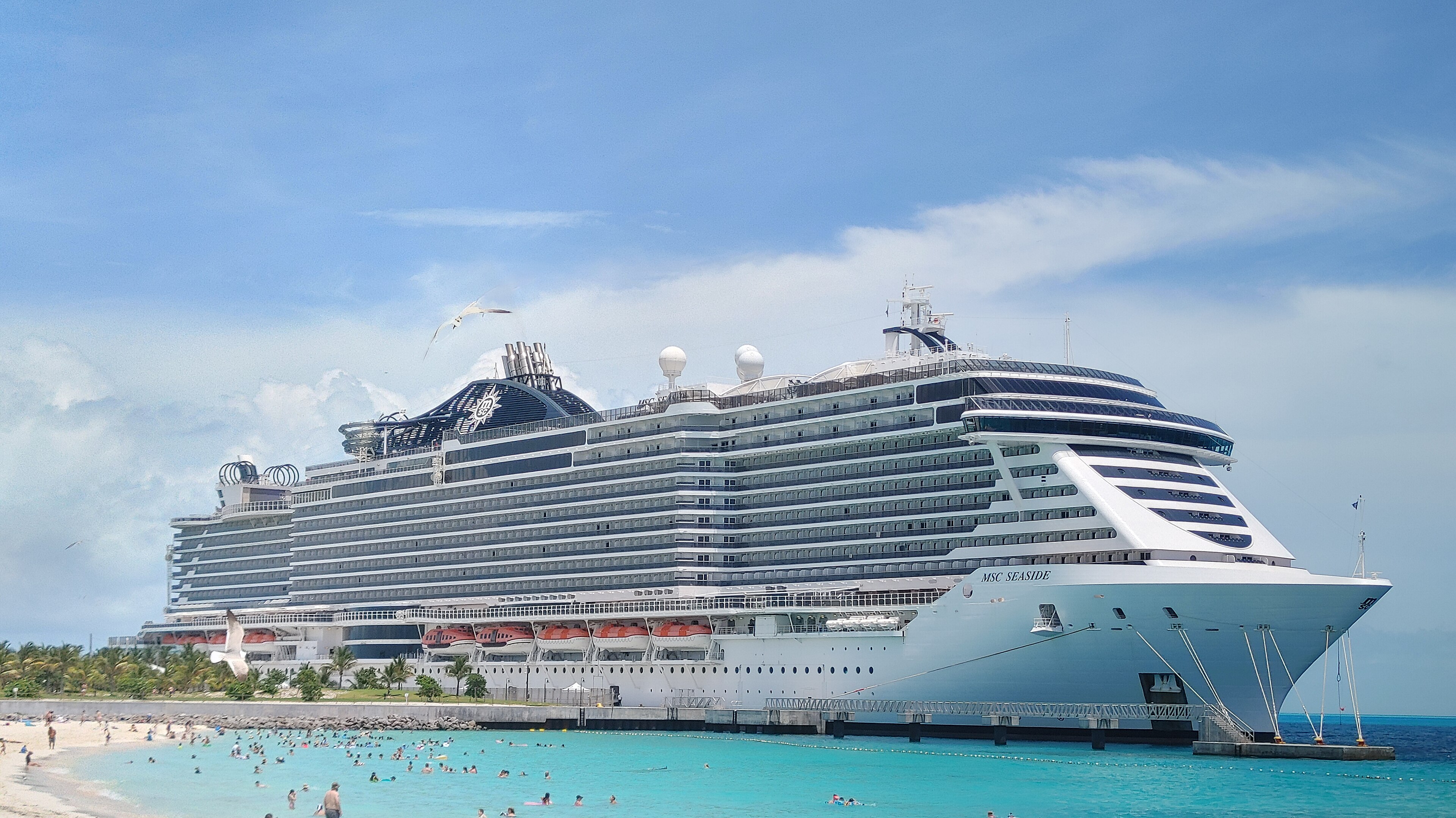 Ein großes Kreuzfahrtschiff liegt im Hafen vor einem Strand mit vielen Menschen im Wasser und an Land.