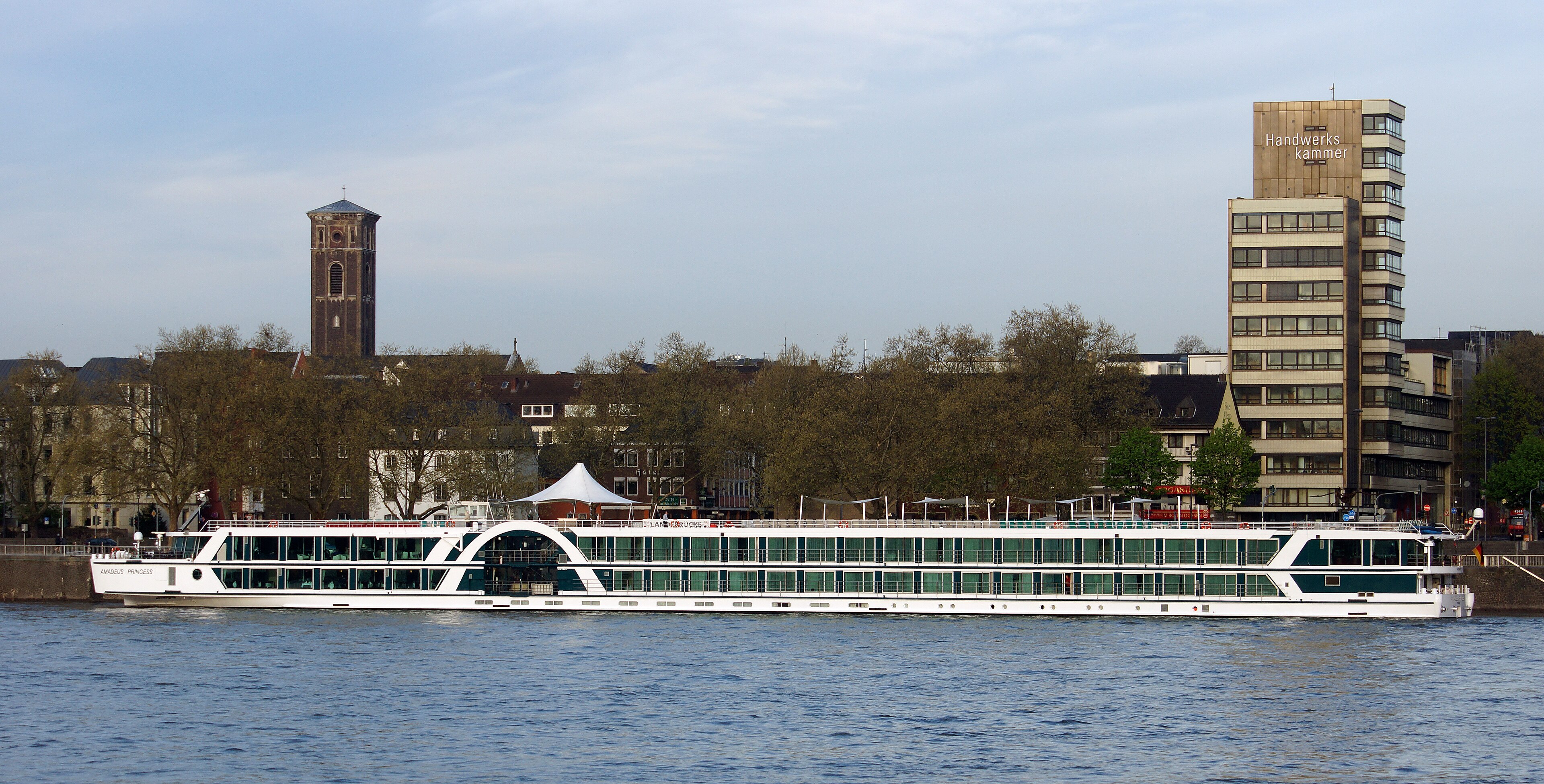 Ein weisses Flusskreuzfahrtschiff mit vielen Fenstern liegt am Ufer einer Stadt. Im Hintergrund sind Haeuser, Baeume und ein Kirchturm zu sehen.