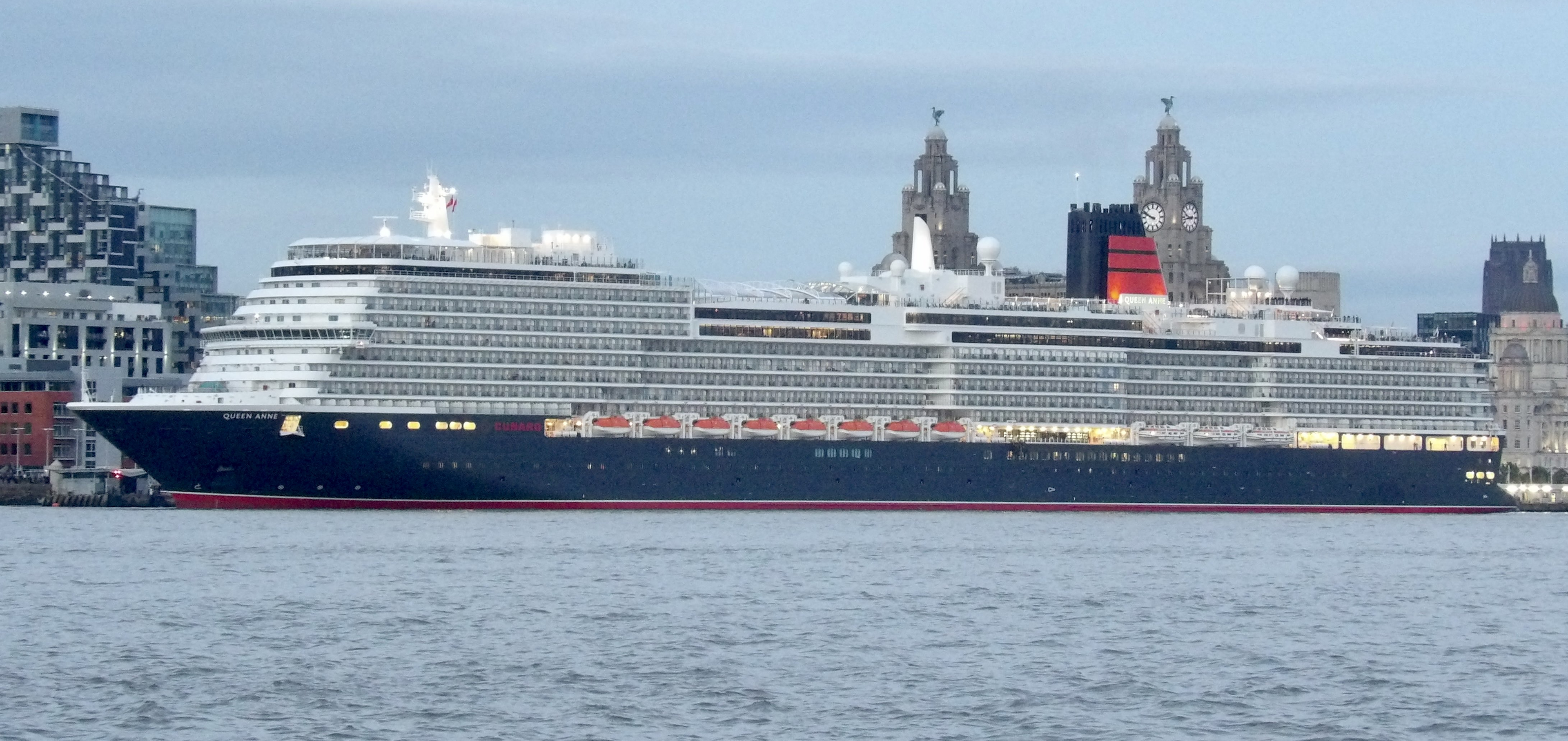 Das Kreuzfahrtschiff Queen Anne liegt im Hafen vor einer Stadtkulisse mit historischen Gebäuden und modernen Hochhäusern.