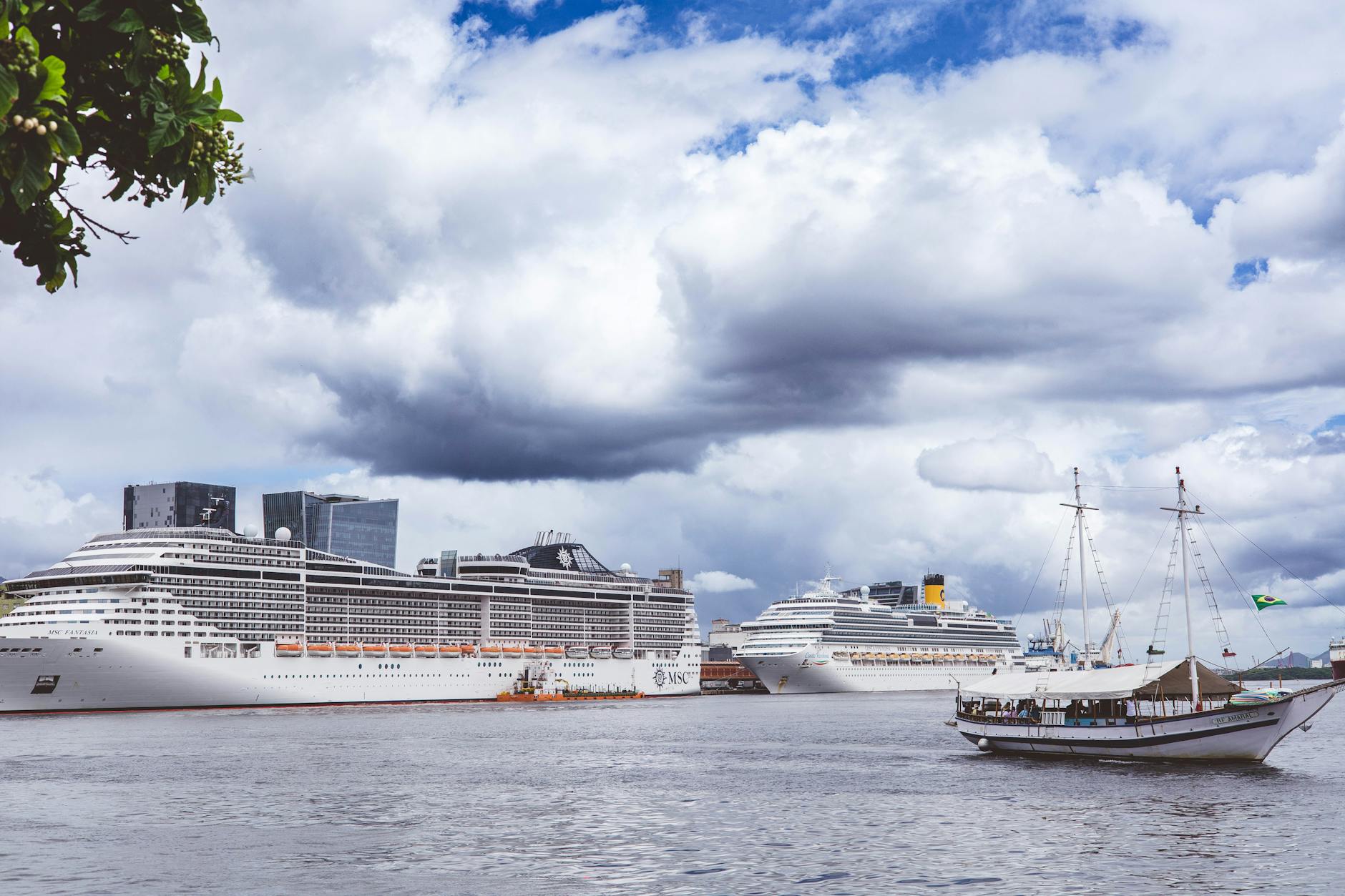 Mehrere Kreuzfahrtschiffe und ein Segelboot liegen im Hafen vor einer Stadtkulisse unter einem bewölkten Himmel.