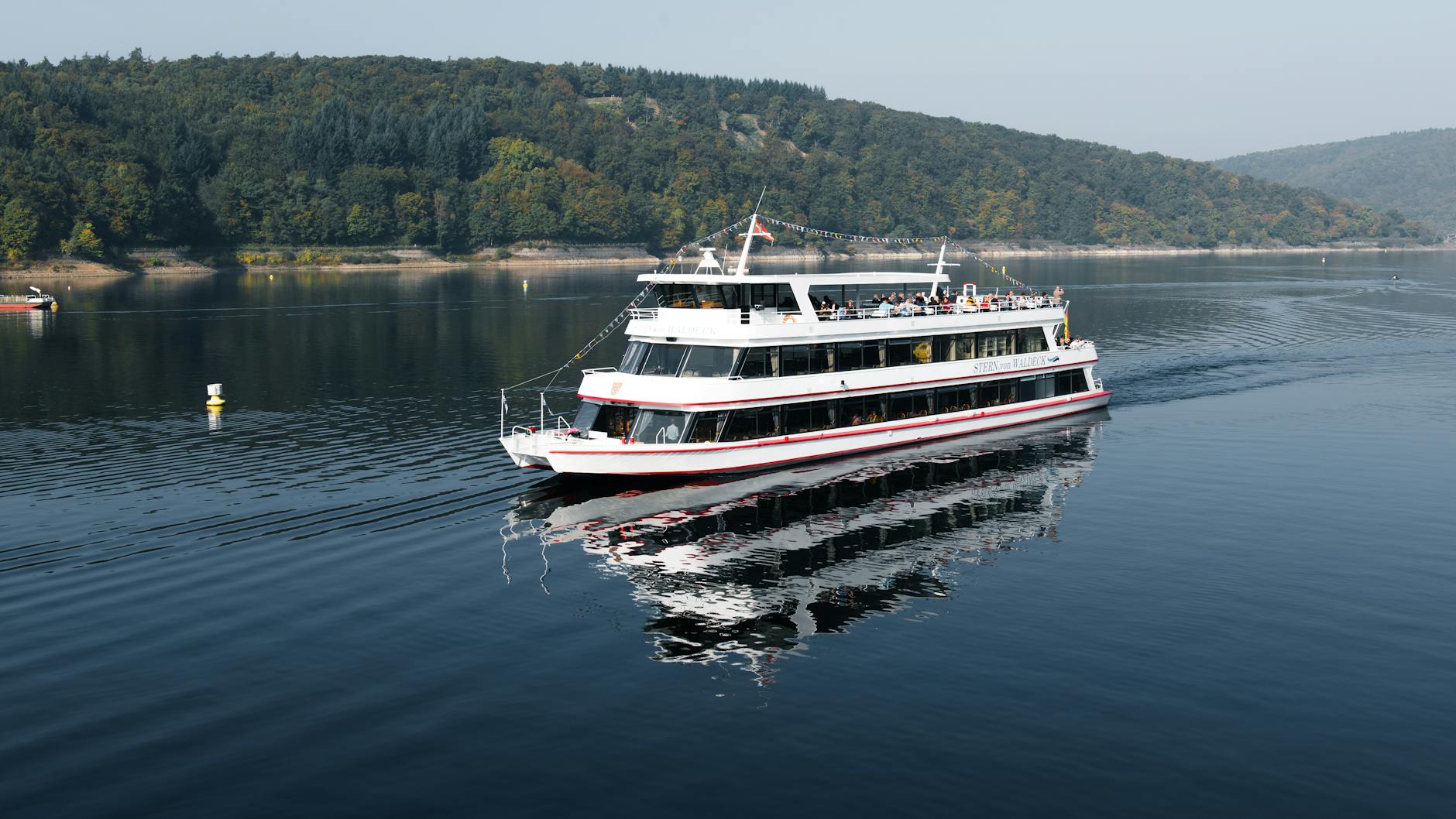 Ein weisses Ausflugsschiff faehrt auf einem ruhigen See, im Hintergrund sind bewaldete Huegel zu sehen.