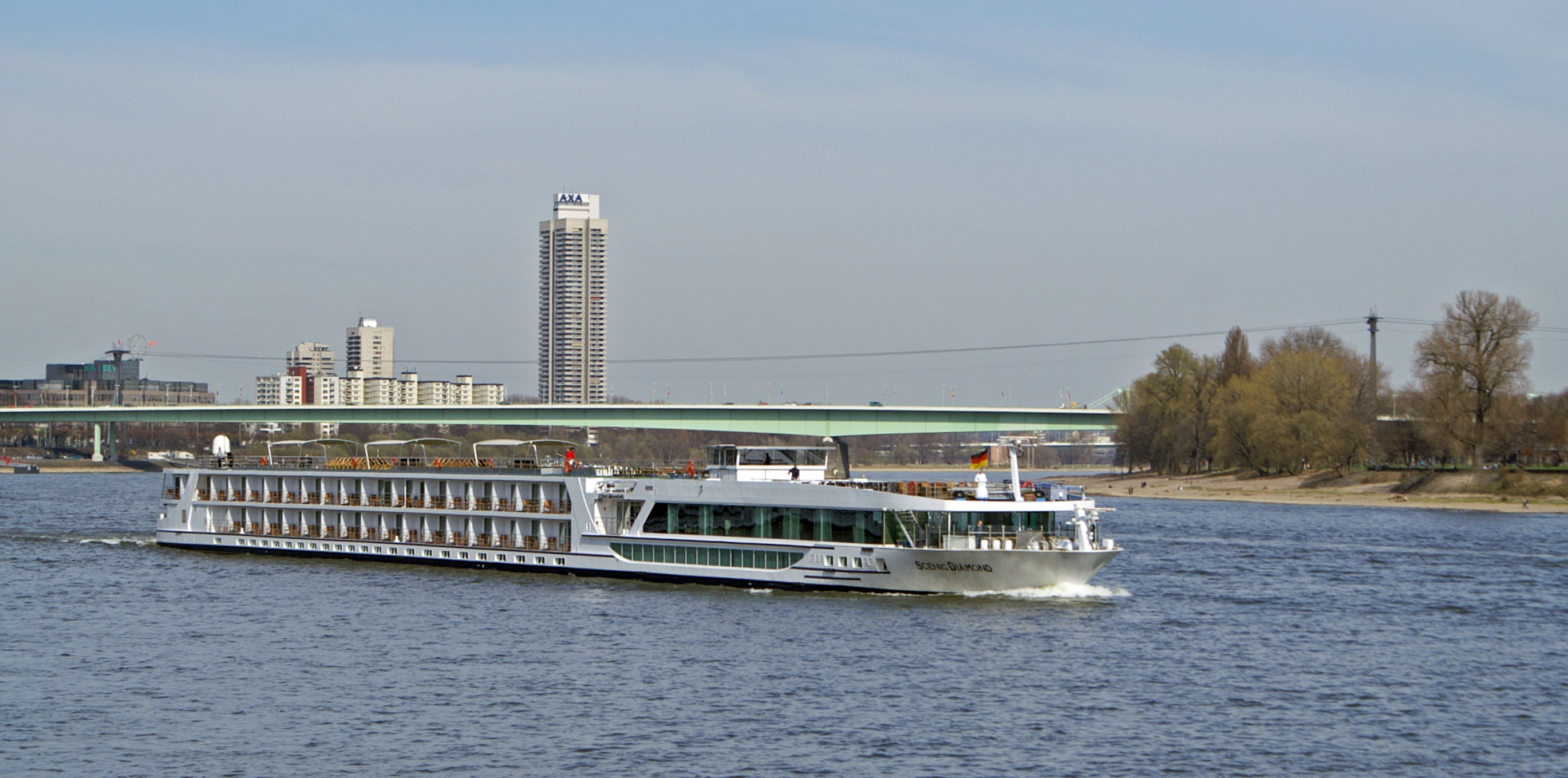 Ein Flusskreuzfahrtschiff faehrt auf einem Fluss an einer Stadt mit einer Bruecke im Hintergrund vorbei.