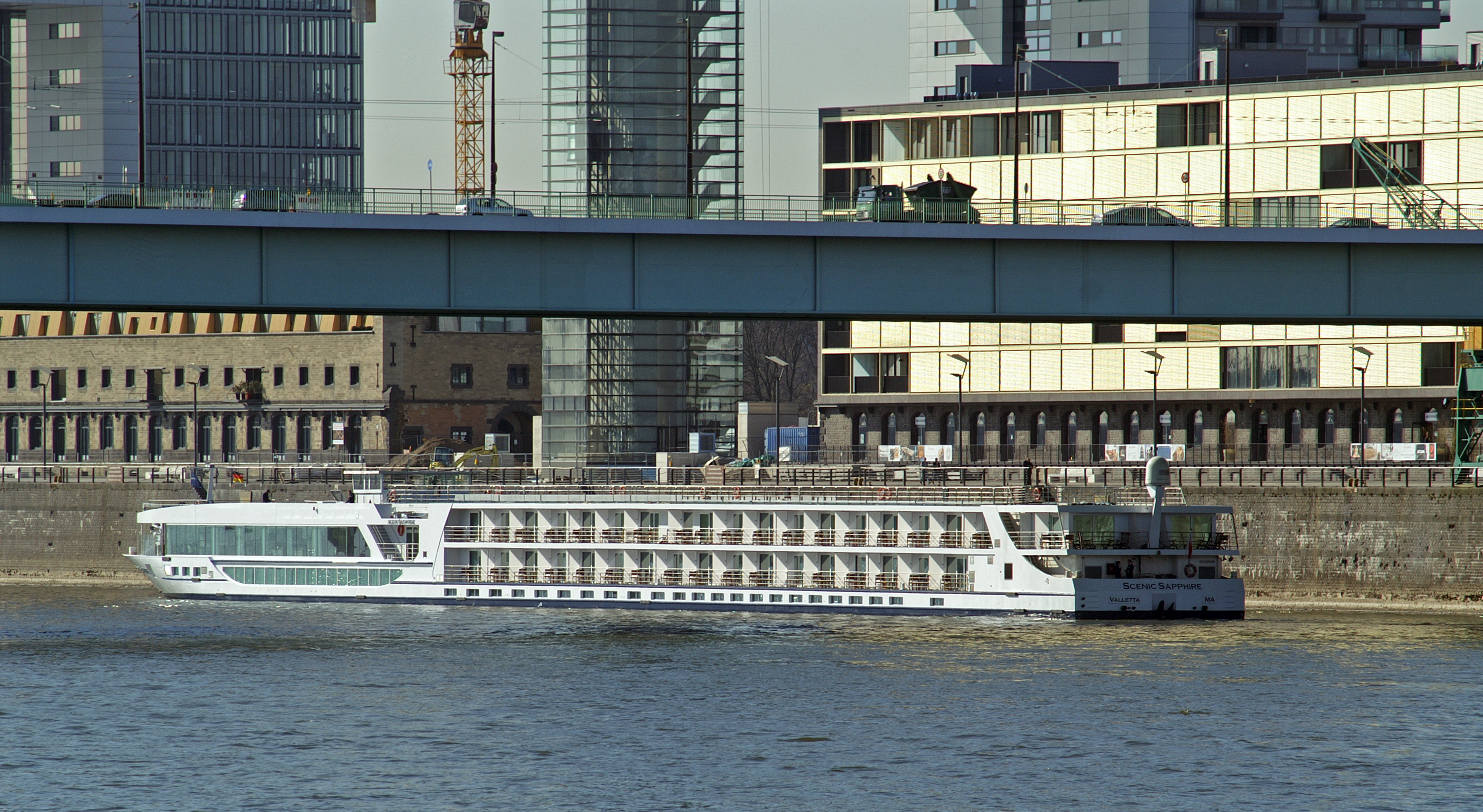 Ein weisses Flusskreuzfahrtschiff faehrt auf einem Fluss, im Hintergrund eine Bruecke und moderne Gebaeude.