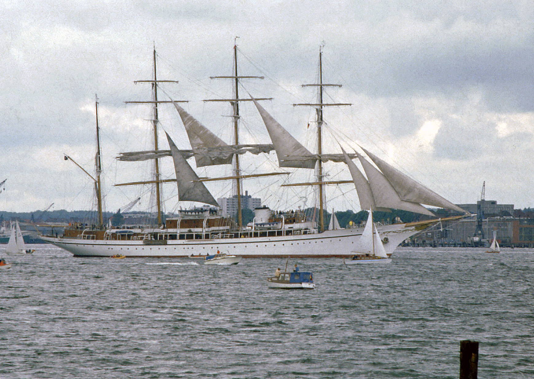 Ein großes weißes Segelschiff mit mehreren Masten und Segeln fährt auf dem Wasser, im Hintergrund ist eine Stadt mit Gebäuden und Kränen zu sehen.