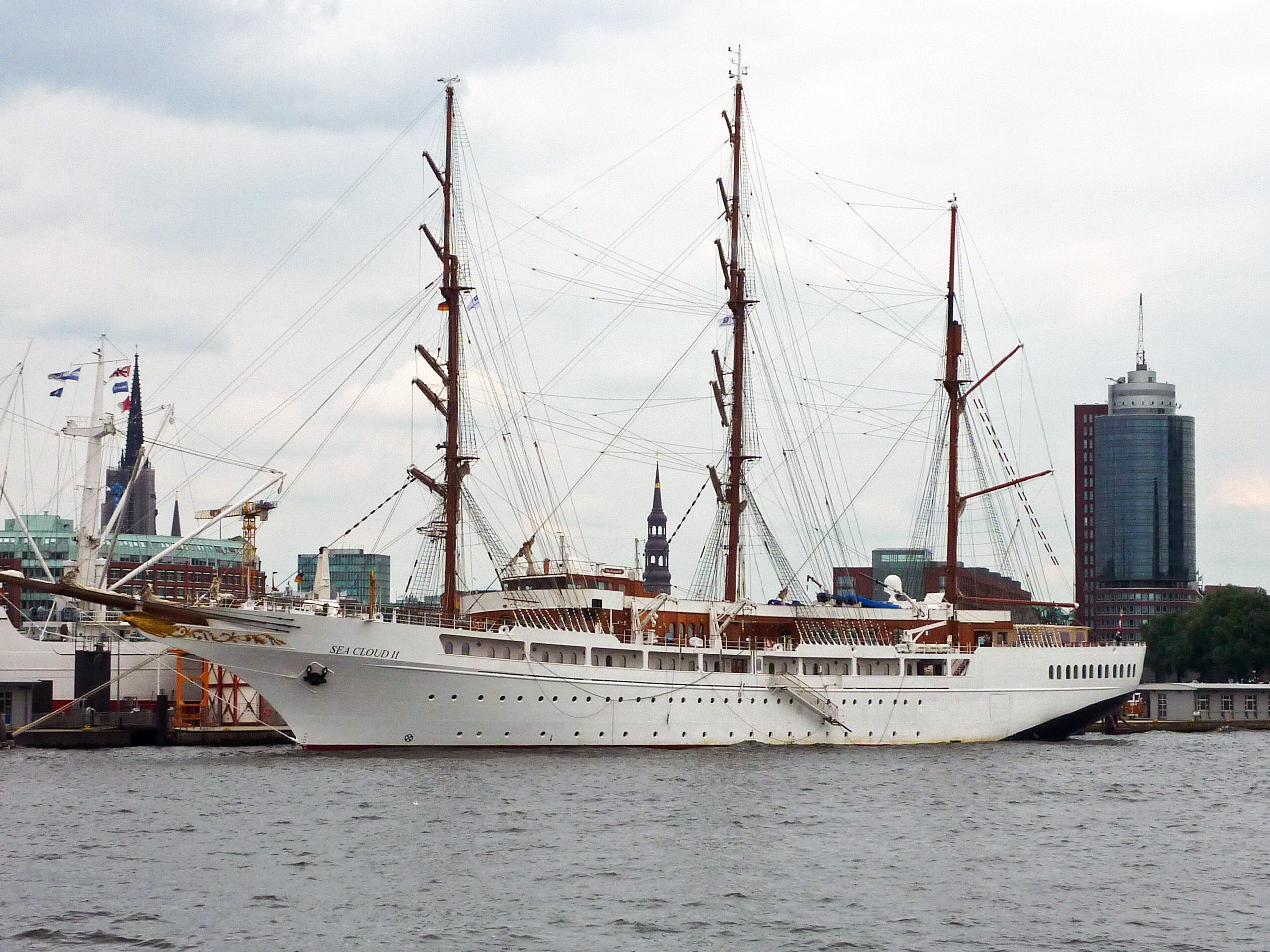 Ein großes weißes Segelschiff mit mehreren Masten liegt im Hafen vor einer Stadtkulisse mit modernen Gebäuden und Kirchtürmen.