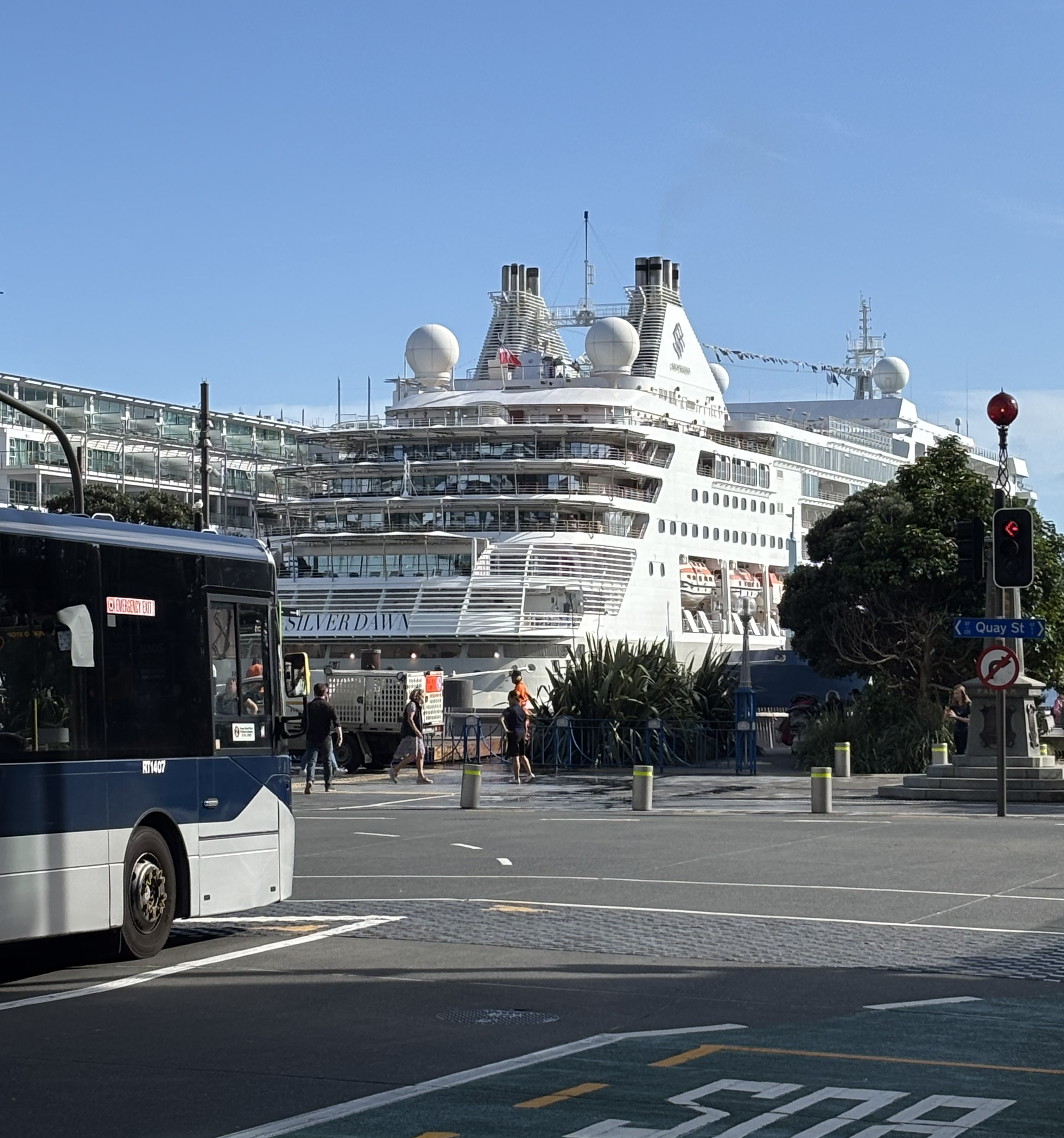 Ein grosses weisses Kreuzfahrtschiff liegt im Hafen, daneben ein blauer Bus und Gebaeude.