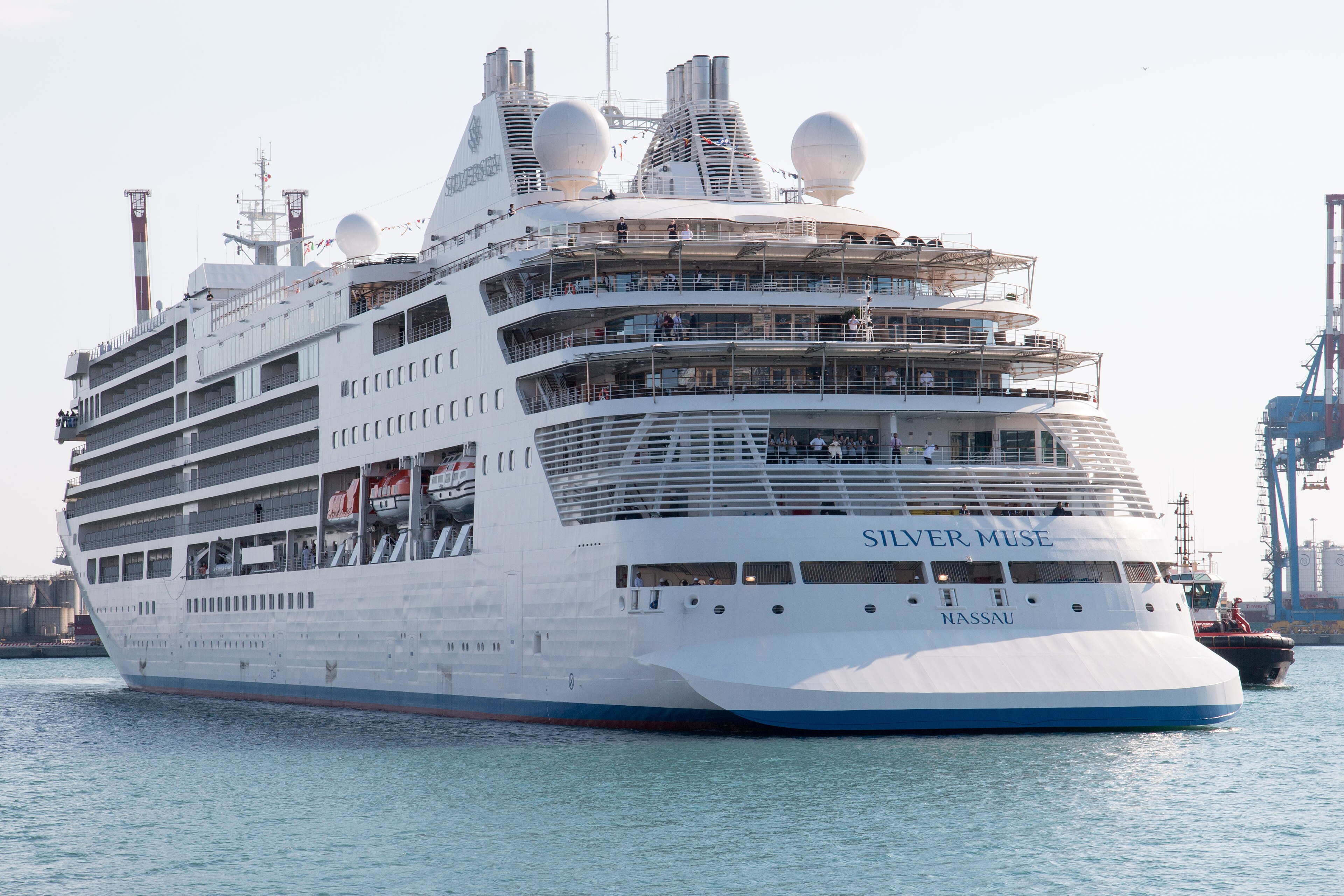 Ein großes weißes Kreuzfahrtschiff namens Silver Muse mit dem Heimathafen Nassau fährt im Wasser, begleitet von einem Schlepper.