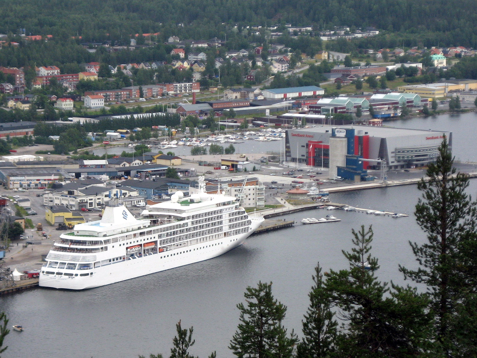 Ein grosses weisses Kreuzfahrtschiff liegt im Hafen neben einer Stadt und einem Sportstadion.