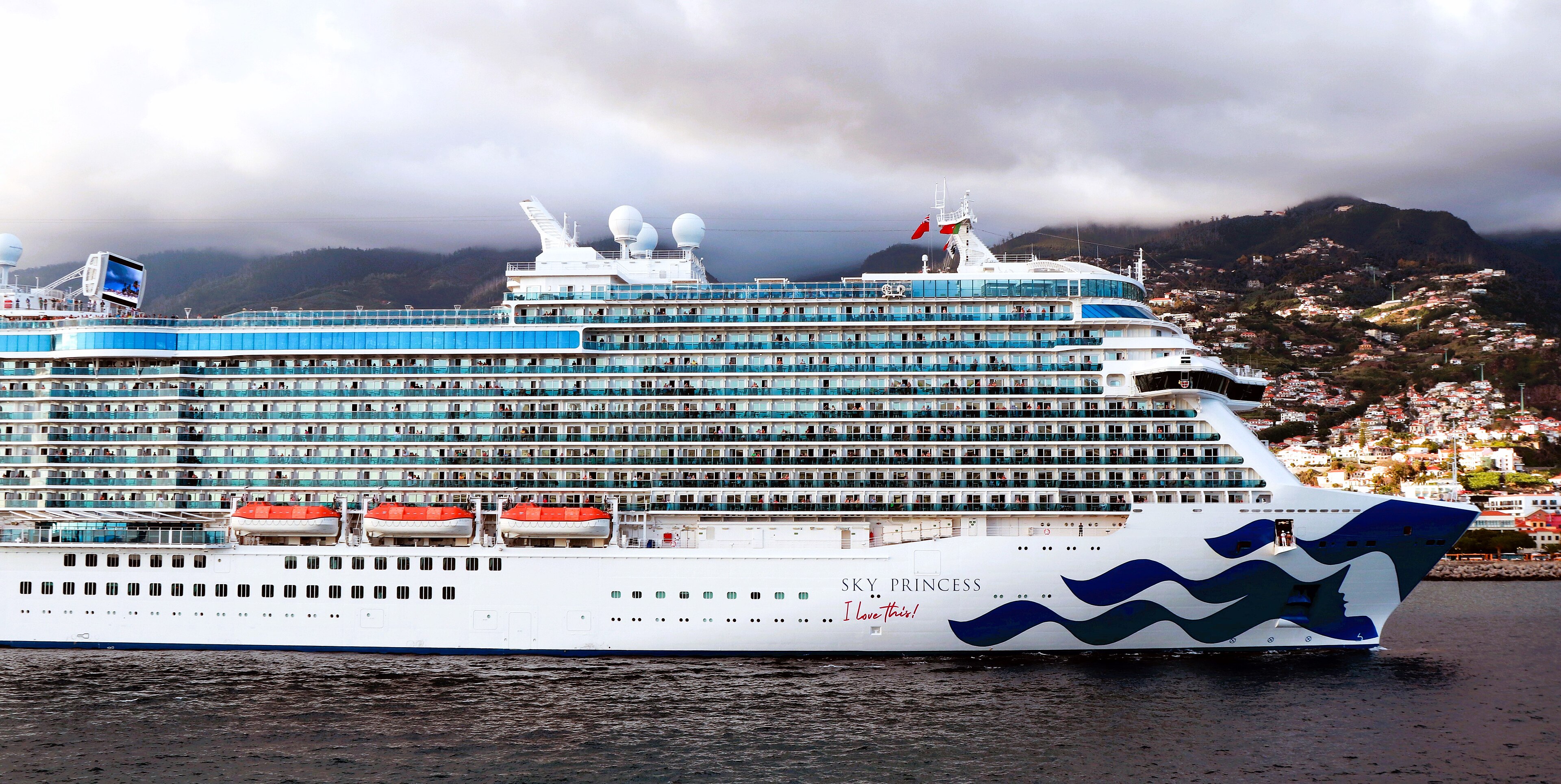 Das Kreuzfahrtschiff Sky Princess liegt im Wasser, im Hintergrund ist eine Stadt an einem Berghang zu sehen.