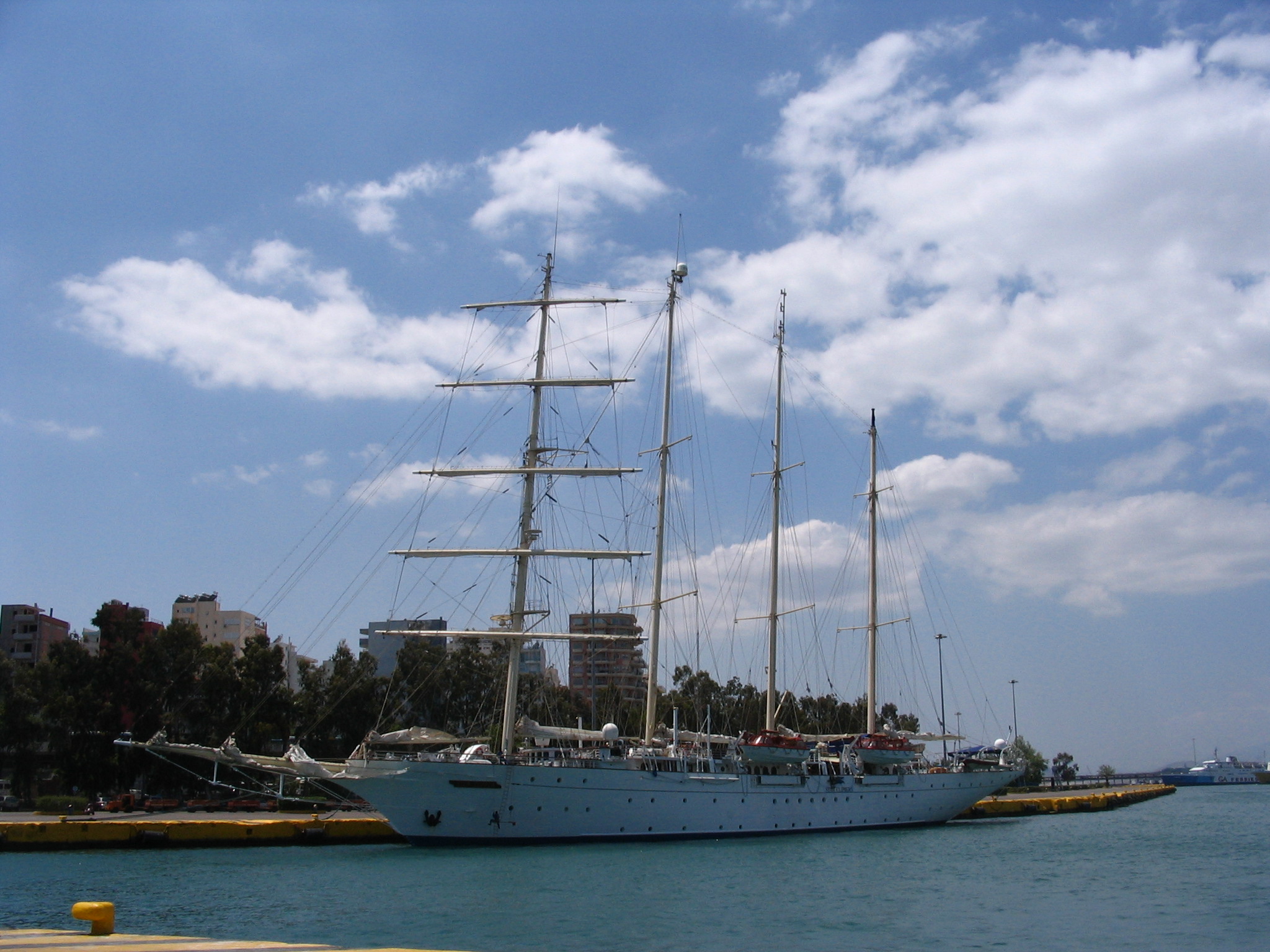 Ein großes Segelschiff mit mehreren Masten liegt im Hafen vor einer Stadtkulisse.