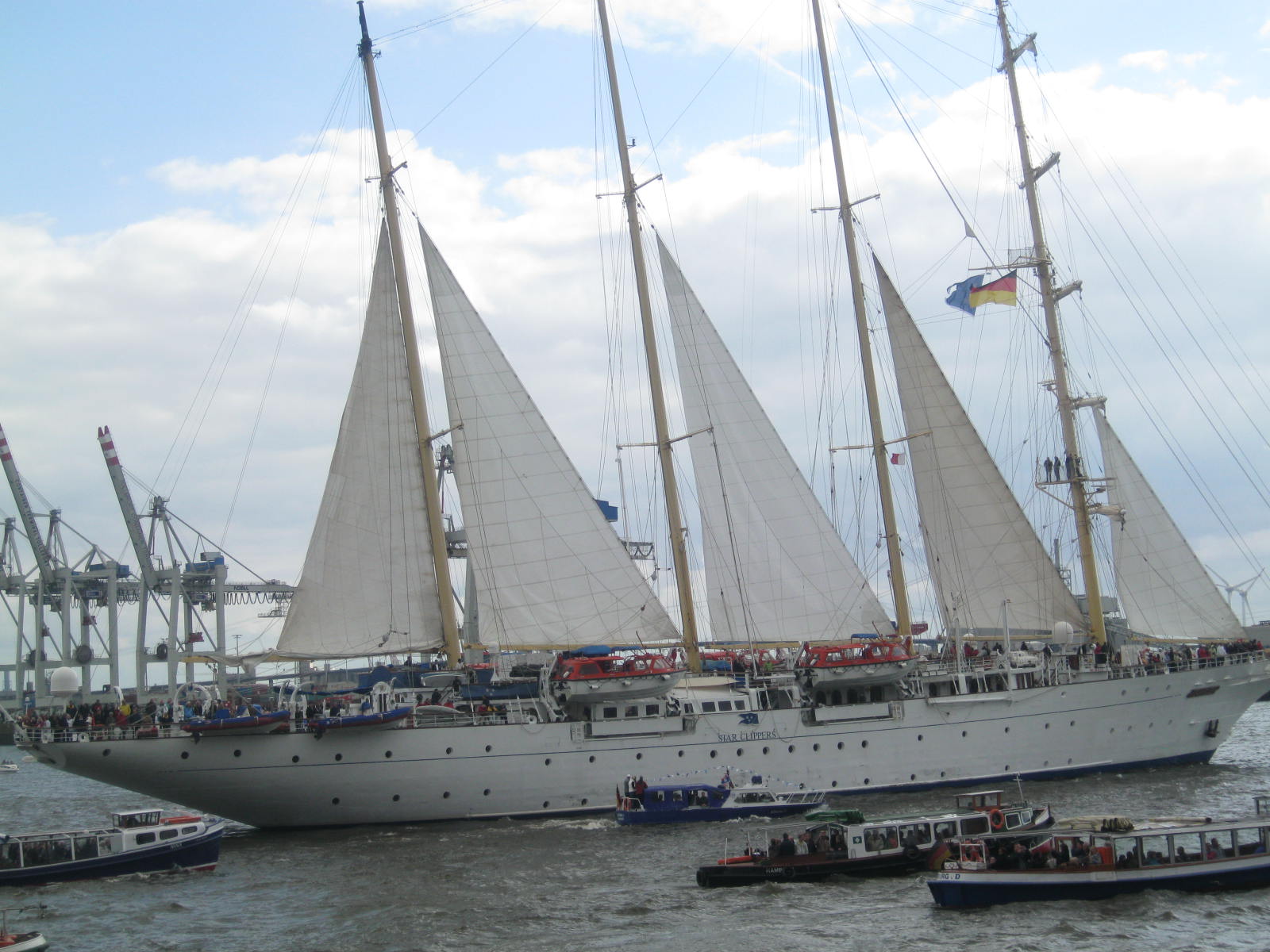 Ein großes weißes Segelschiff mit vier Masten fährt auf dem Wasser, im Hintergrund sind große Hafenkrane zu sehen.
